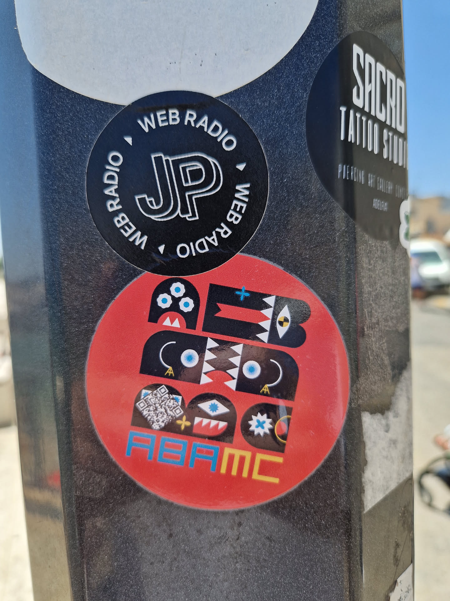 A red circular sticker features an abstract design composed of black shapes, blue eyes, and geometric patterns.  At the bottom, the text "ABAMC" is visible. A small QR code is also included within the design.