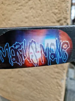 A sticker featuring the name Marilyn Manson in a stylized, dark blue font against a backdrop of vibrant, blurry colors reminiscent of a fiery sunset or explosion. The sticker is affixed to a dark gray metal bar. 
