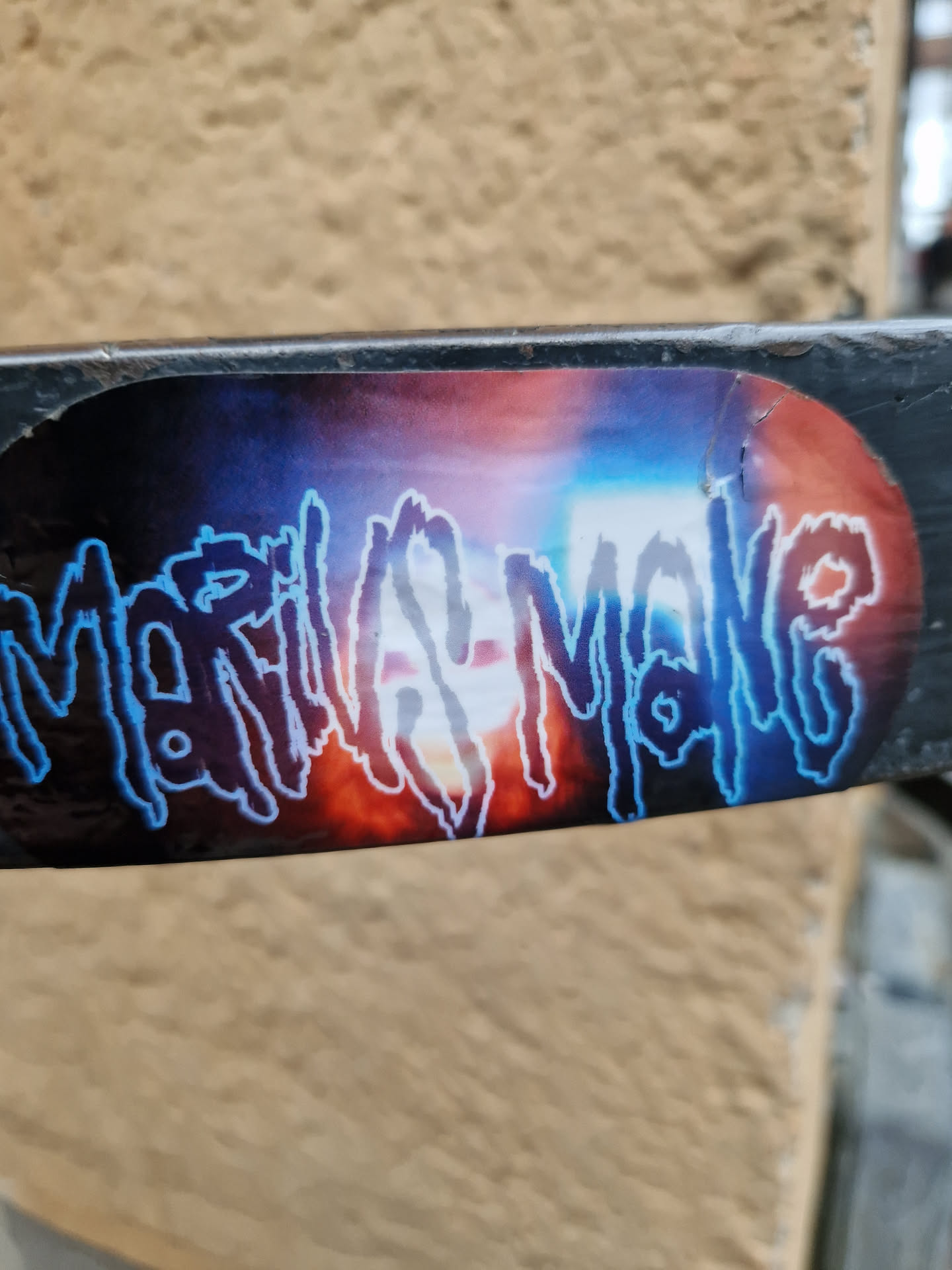 A sticker featuring the name Marilyn Manson in a stylized, dark blue font against a backdrop of vibrant, blurry colors reminiscent of a fiery sunset or explosion. The sticker is affixed to a dark gray metal bar. 