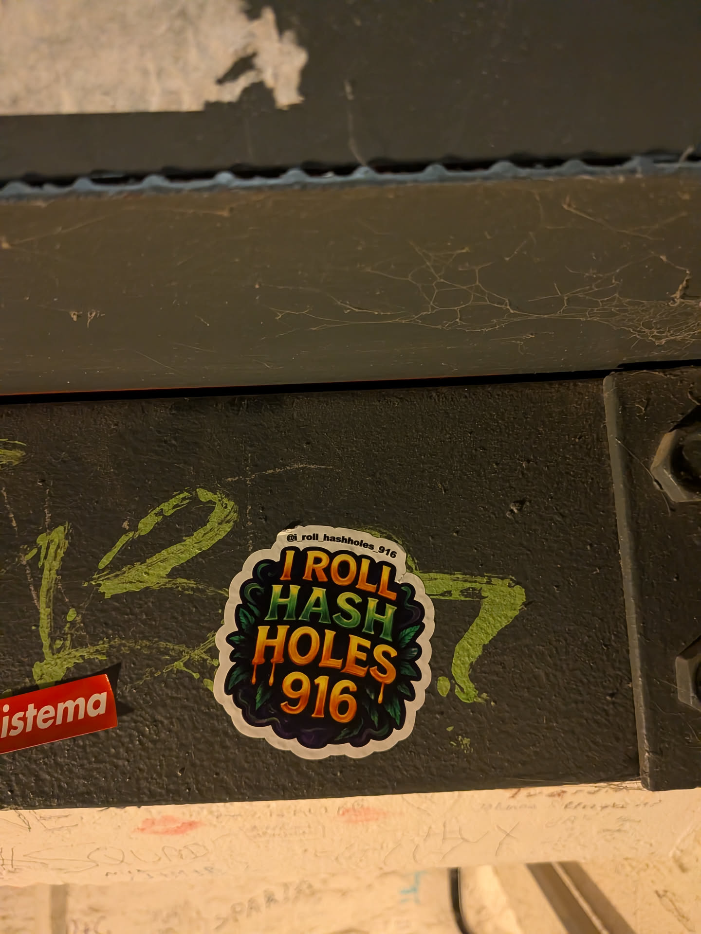 A die-cut sticker with a white border featuring vibrant, stylized text set against a dark backdrop framed by dark green cannabis leaves. The text reads 'I ROLL' in warm orange and yellow, 'HASH' in bright green tones, and 'HOLES 916' in dripping orange and golden hues. Above the main graphic, an Instagram handle '@i_roll_hashholes_916' is printed in small black text along the upper edge.