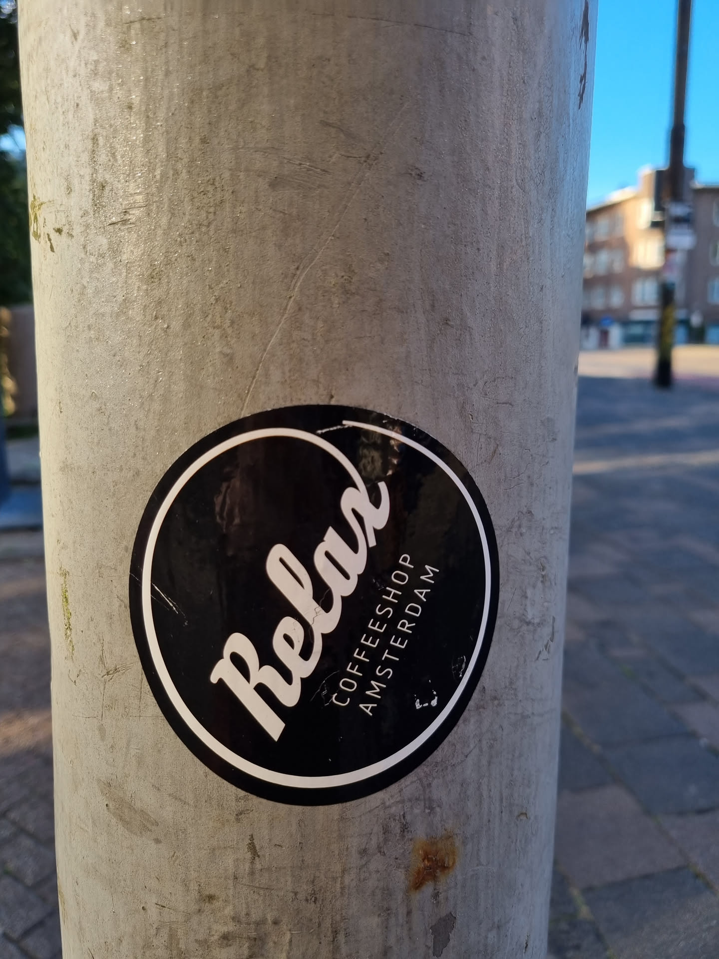 A black and white circular sticker with the word 'Relax' in a stylized font.  Below the word, the text 'COFFEESHOP AMSTERDAM' is printed in a smaller font. The sticker is affixed to a weathered grey pole.