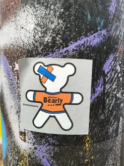 A sticker featuring a cartoon bear wearing an orange shirt with the words 'Mister Bearly' on it. The bear has a small bandage on its head. The sticker is on a dark surface with other graffiti.