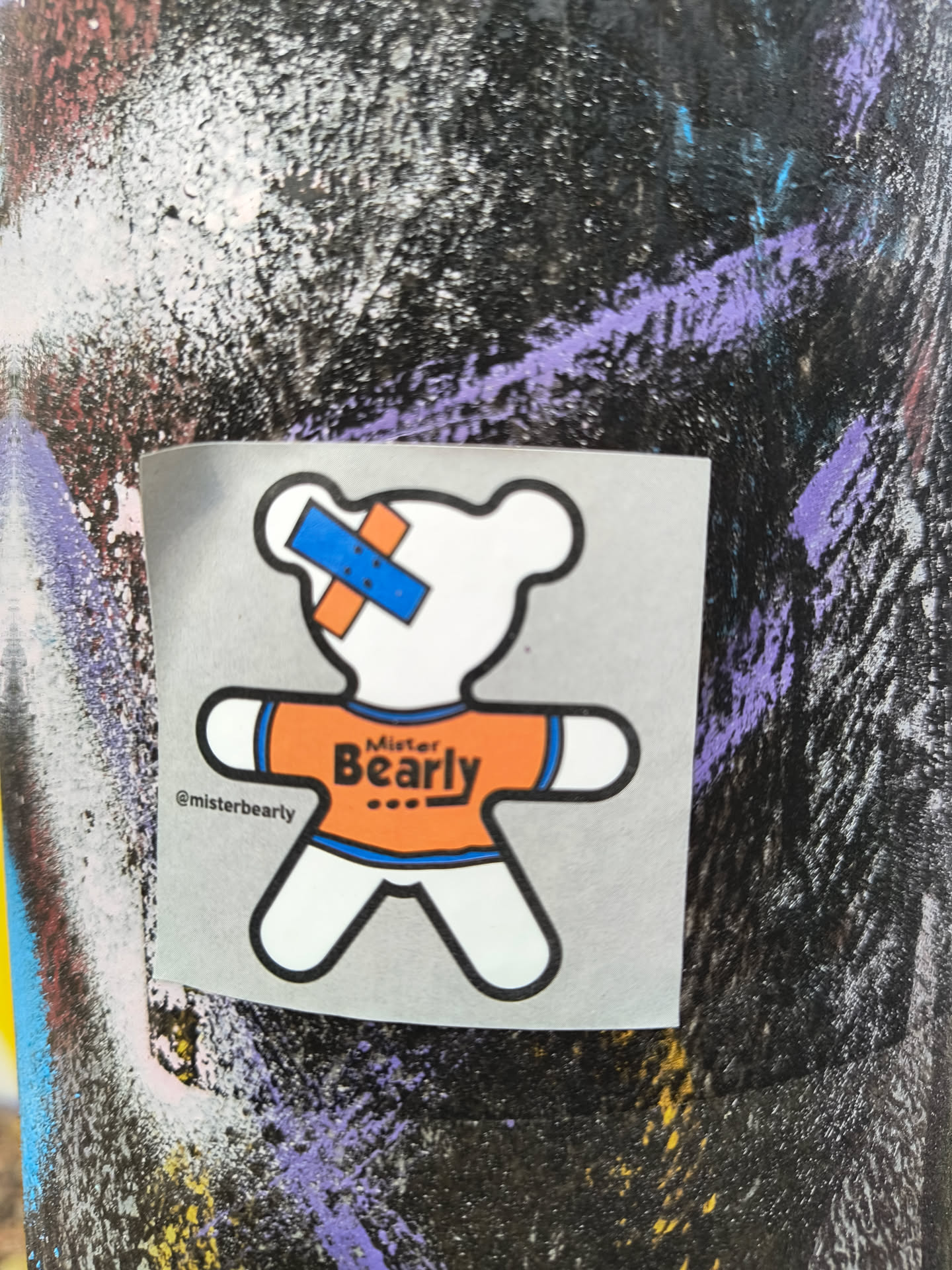 A sticker featuring a cartoon bear wearing an orange shirt with the words 'Mister Bearly' on it. The bear has a small bandage on its head. The sticker is on a dark surface with other graffiti.