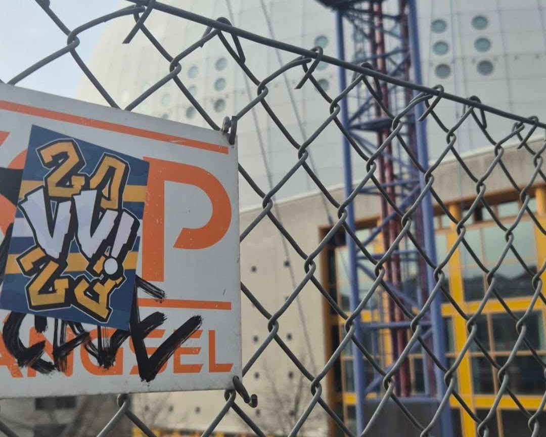 A rectangular sticker with stylized 'VV' lettering and the numbers '24' and '23' is pasted on an orange and white 'P' parking sign. The sticker is in a navy blue square with an orange border. The word 'ANGEL' is written below with black marker with a tick.