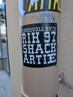 Street sticker A square-ish black sticker adhered to a beige metal post. It features bold, stylized white lettering in a vintage Western or circus-style slab-serif font. The top line reads 'TJENSVOLL BOYS' in smaller caps, followed by three lines of larger bold text: 'RIK 97', 'SHACK', and 'ARTIE'.