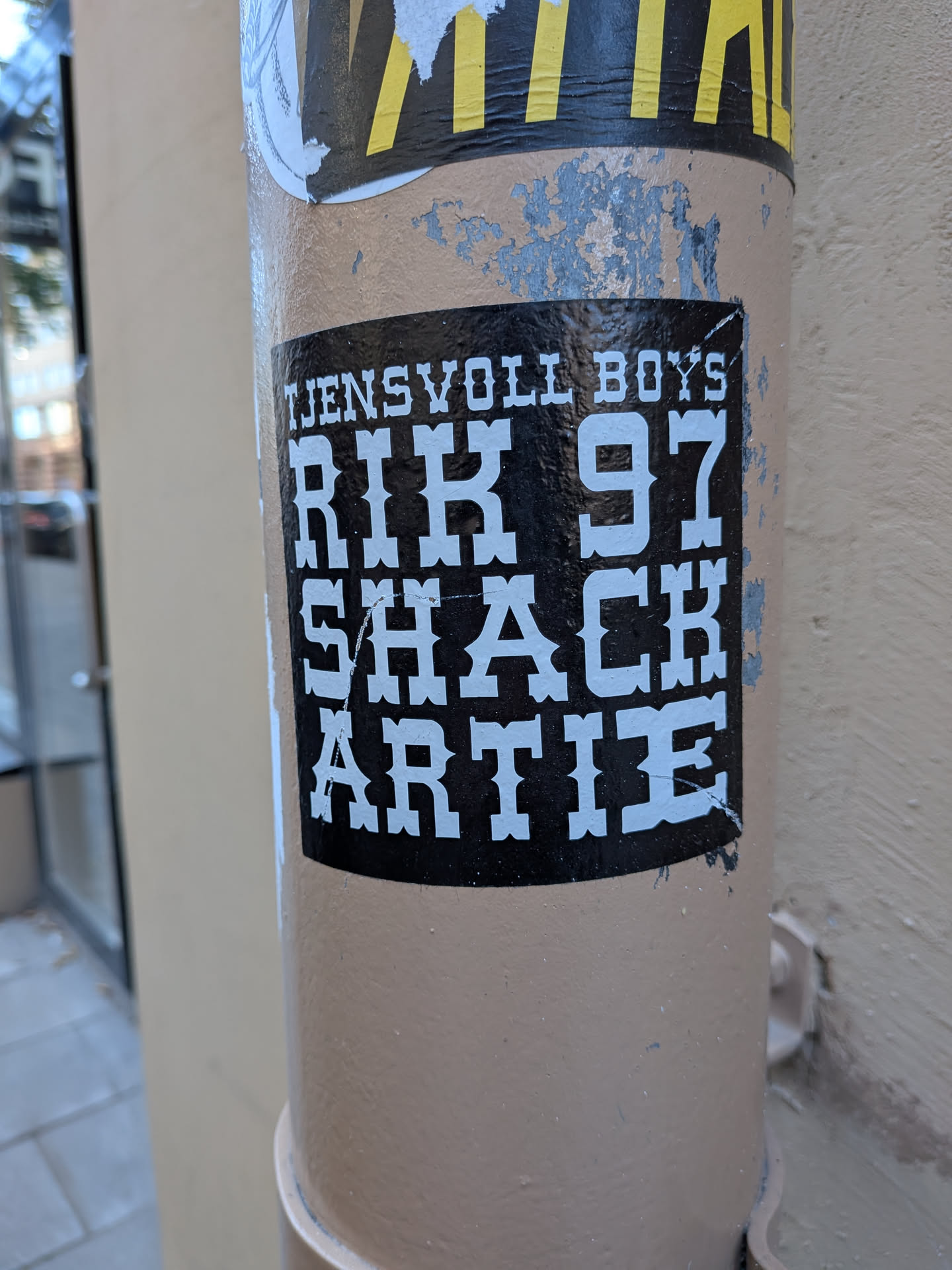 A square-ish black sticker adhered to a beige metal post. It features bold, stylized white lettering in a vintage Western or circus-style slab-serif font. The top line reads 'TJENSVOLL BOYS' in smaller caps, followed by three lines of larger bold text: 'RIK 97', 'SHACK', and 'ARTIE'.