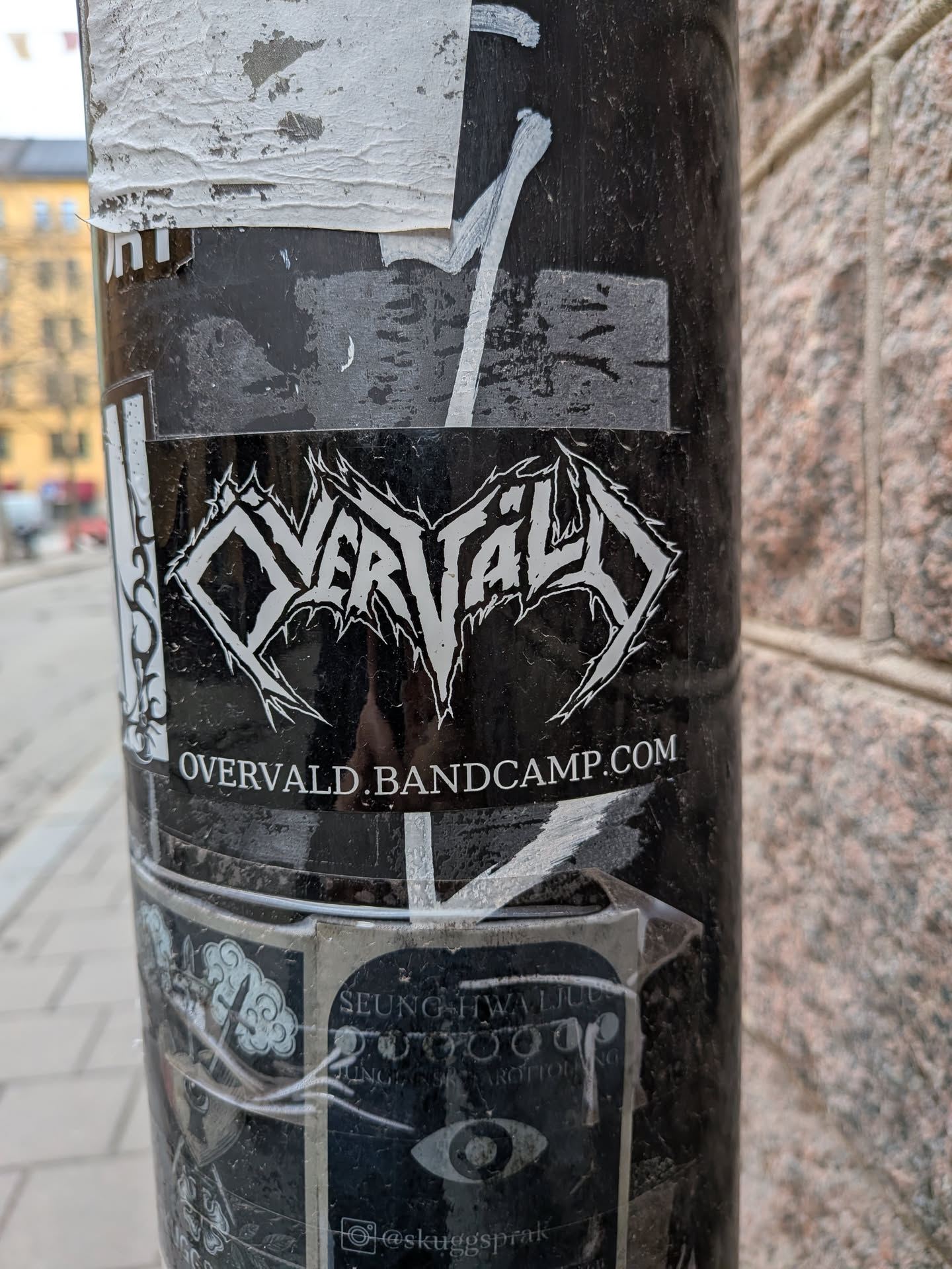 A rectangular black sticker featuring the band logo 'OVERVALD' in a prominent, highly stylized white font. The letters are sharp, jagged, and spiky, characteristic of black or death metal typography. Below the logo, in a smaller, clear white sans-serif font, is the website 'OVERVALD.BANDCAMP.COM'. The sticker appears slightly worn and scratched, affixed to a dark pole, with some minor peeling at the edges.