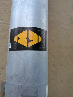 A rectangular black sticker adhered to a vertical metal pole. In the center is a bold, mustard-yellow geometric logo forming a faceted hexagon or stylized eye shape. The emblem is divided into segmented angular parts separated by fine black negative space. The sticker has a slightly arched top edge and shows a small tear on its left margin.