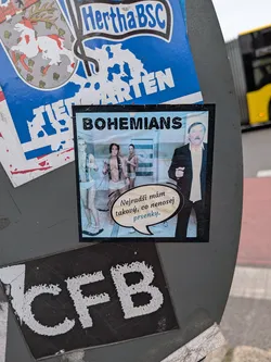 Street sticker A square photomontage sticker with a thin black border. Across the top, the word 'BOHEMIANS' is printed in bold black capital letters. On the right side, legendary Czech footballer Antonín Panenka is shown wearing a dark blazer over a cream-colored collared shirt. To the left is a scene set in a tiled shower or locker room where three women wearing white briefs and suspenders are visible, with a Bohemians flag hanging on the wall. A speech bubble pointing from Panenka contains Czech text with the last word styled in light blue.