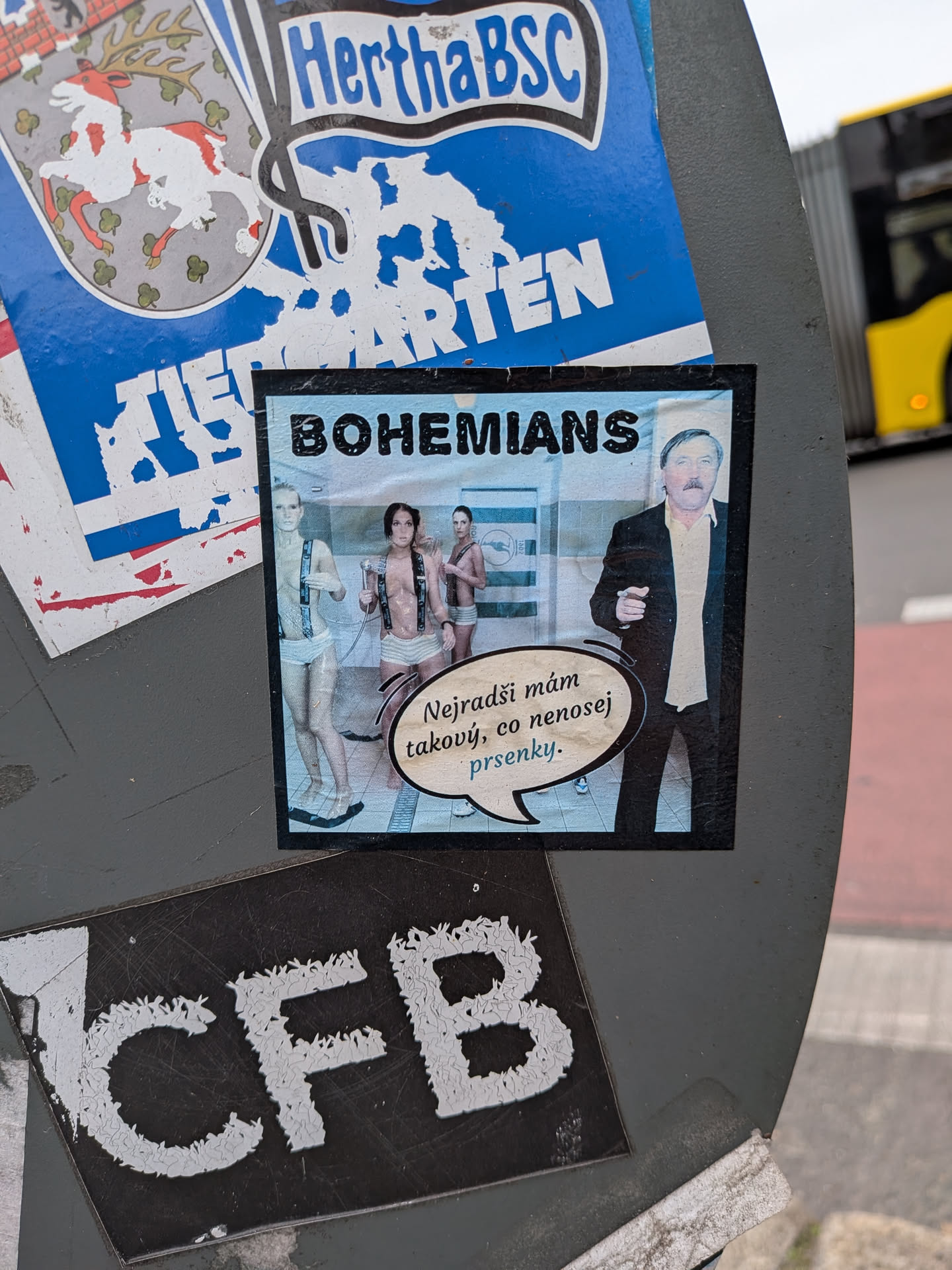 A square photomontage sticker with a thin black border. Across the top, the word 'BOHEMIANS' is printed in bold black capital letters. On the right side, legendary Czech footballer Antonín Panenka is shown wearing a dark blazer over a cream-colored collared shirt. To the left is a scene set in a tiled shower or locker room where three women wearing white briefs and suspenders are visible, with a Bohemians flag hanging on the wall. A speech bubble pointing from Panenka contains Czech text with the last word styled in light blue.