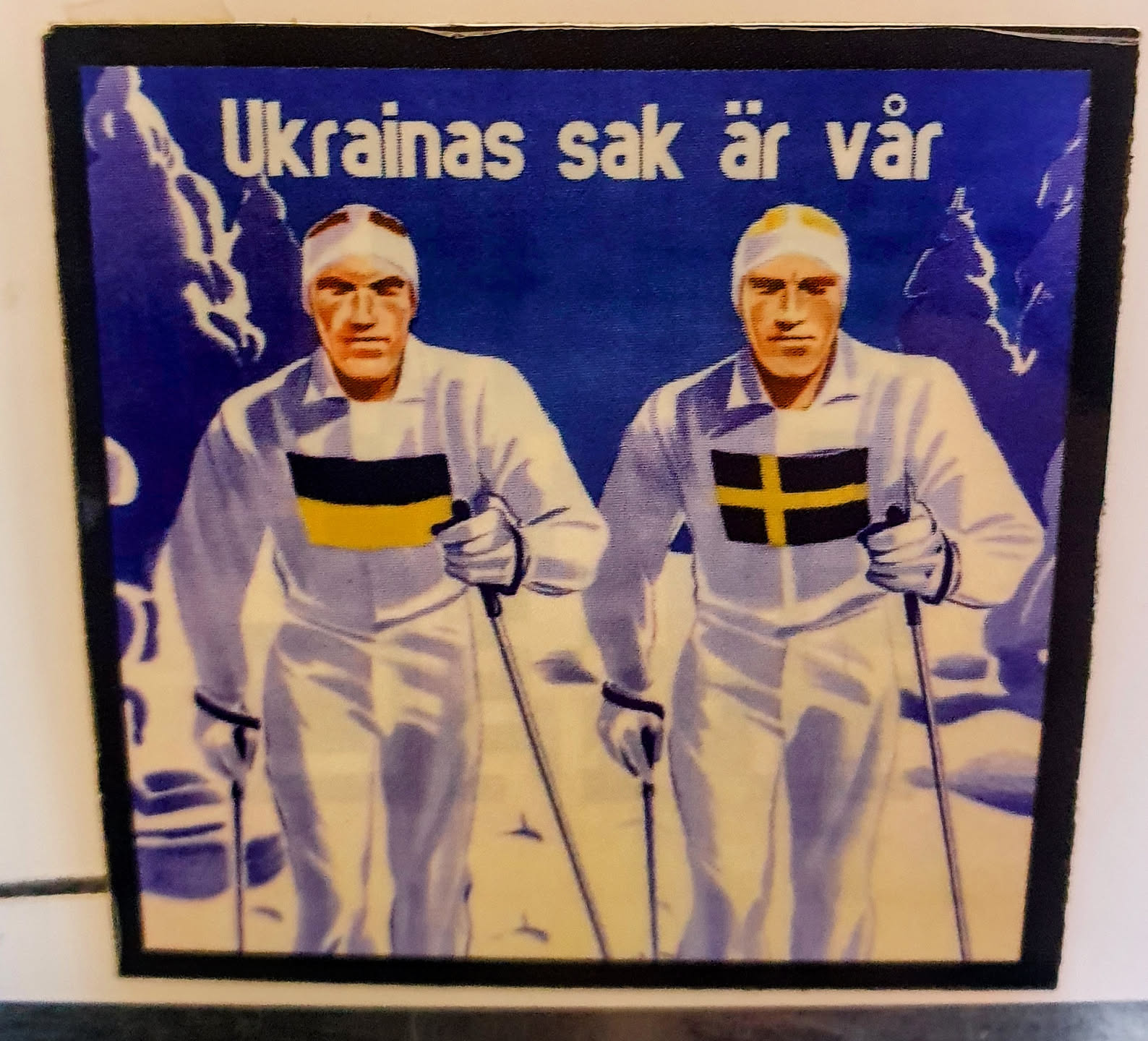 A sticker depicting two cross-country skiers, one with a Ukrainian flag and another with a Swedish flag on their chests. The background is a snowy landscape. The text on top translates to "Ukraine's cause is our cause" in Swedish.