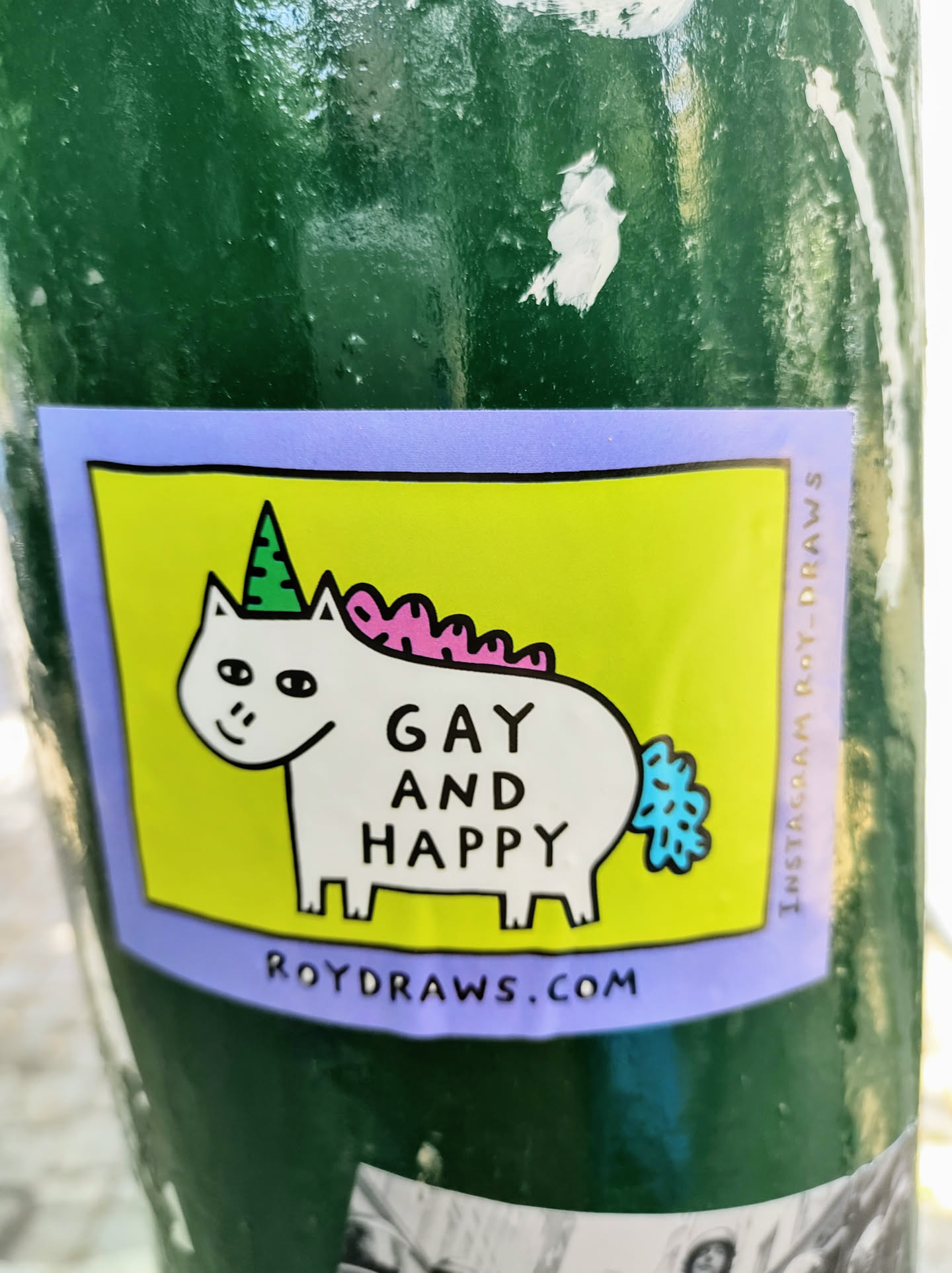 A sticker featuring a cartoon unicorn with a party hat, wearing a pink mane and a blue tail. The text "Gay and Happy" is written on the sticker, along with the website roydraws.com. 