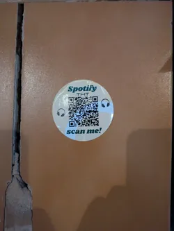 The sticker is round and has a light background. It says "Spotify THT" and has a QR code in the center with headphone icons on either side. The text "scan me!" is at the bottom of the sticker.