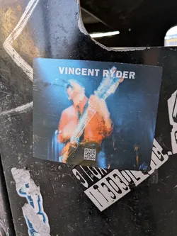 The sticker features a photograph of a musician, identified as Vincent Ryder, playing a guitar on stage. The musician is wearing an orange shirt and is illuminated by blue stage lighting, creating a dynamic and energetic atmosphere. A QR code is visible in the lower left corner of the sticker.