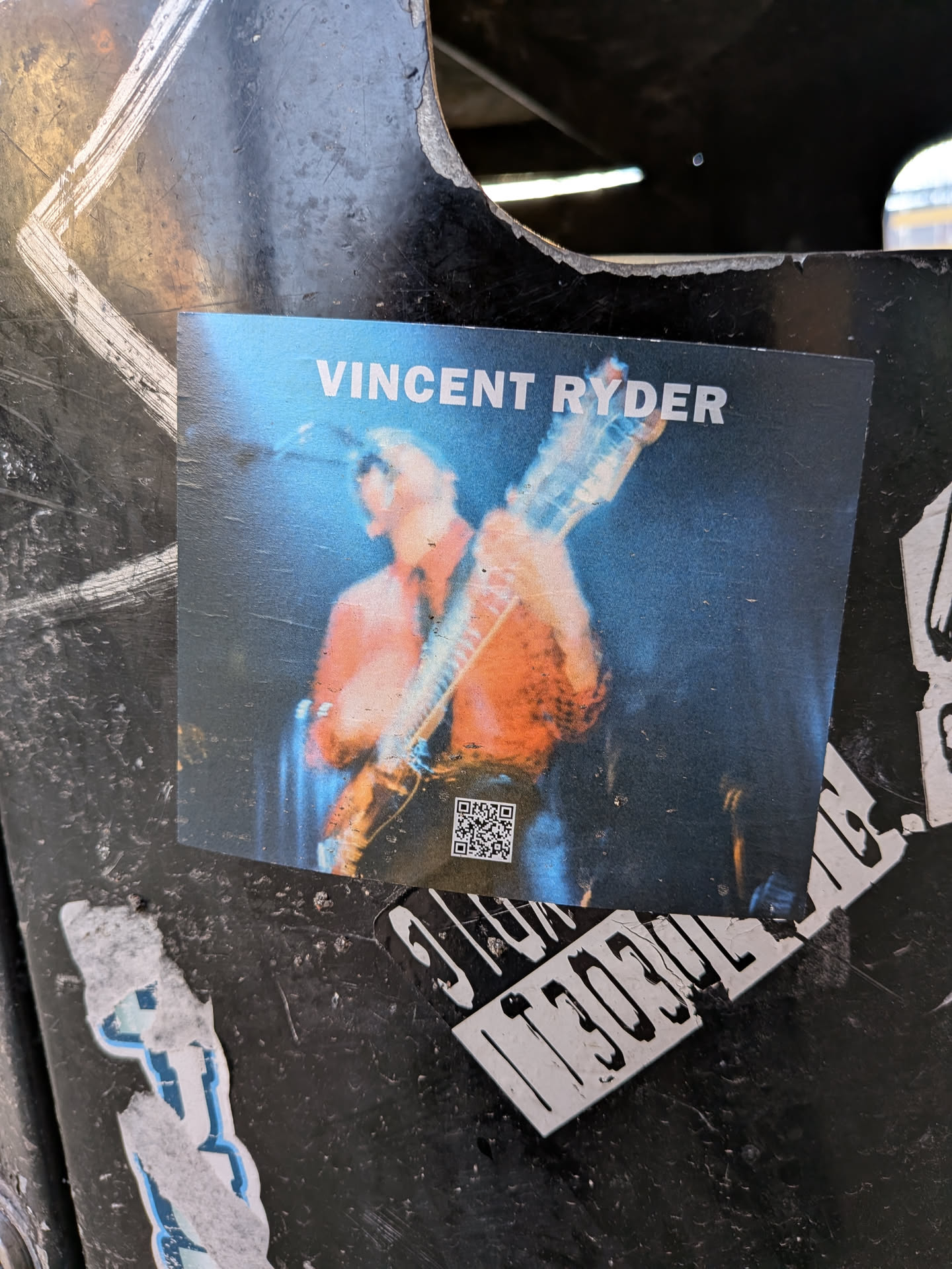 The sticker features a photograph of a musician, identified as Vincent Ryder, playing a guitar on stage. The musician is wearing an orange shirt and is illuminated by blue stage lighting, creating a dynamic and energetic atmosphere. A QR code is visible in the lower left corner of the sticker.