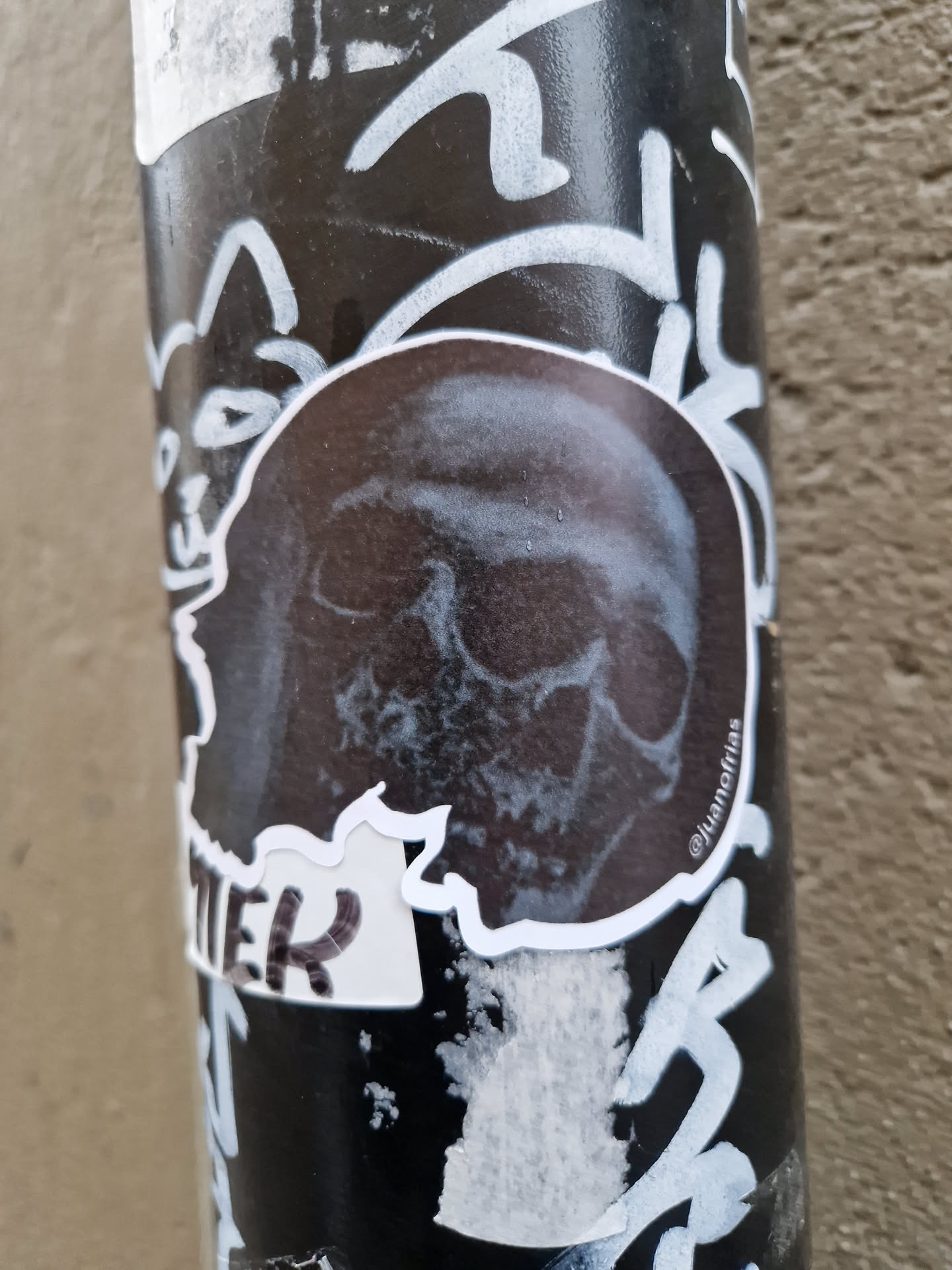 A black and white sticker of a skull with some text on the bottom. The sticker is placed on a pole that is covered in other stickers and graffiti.