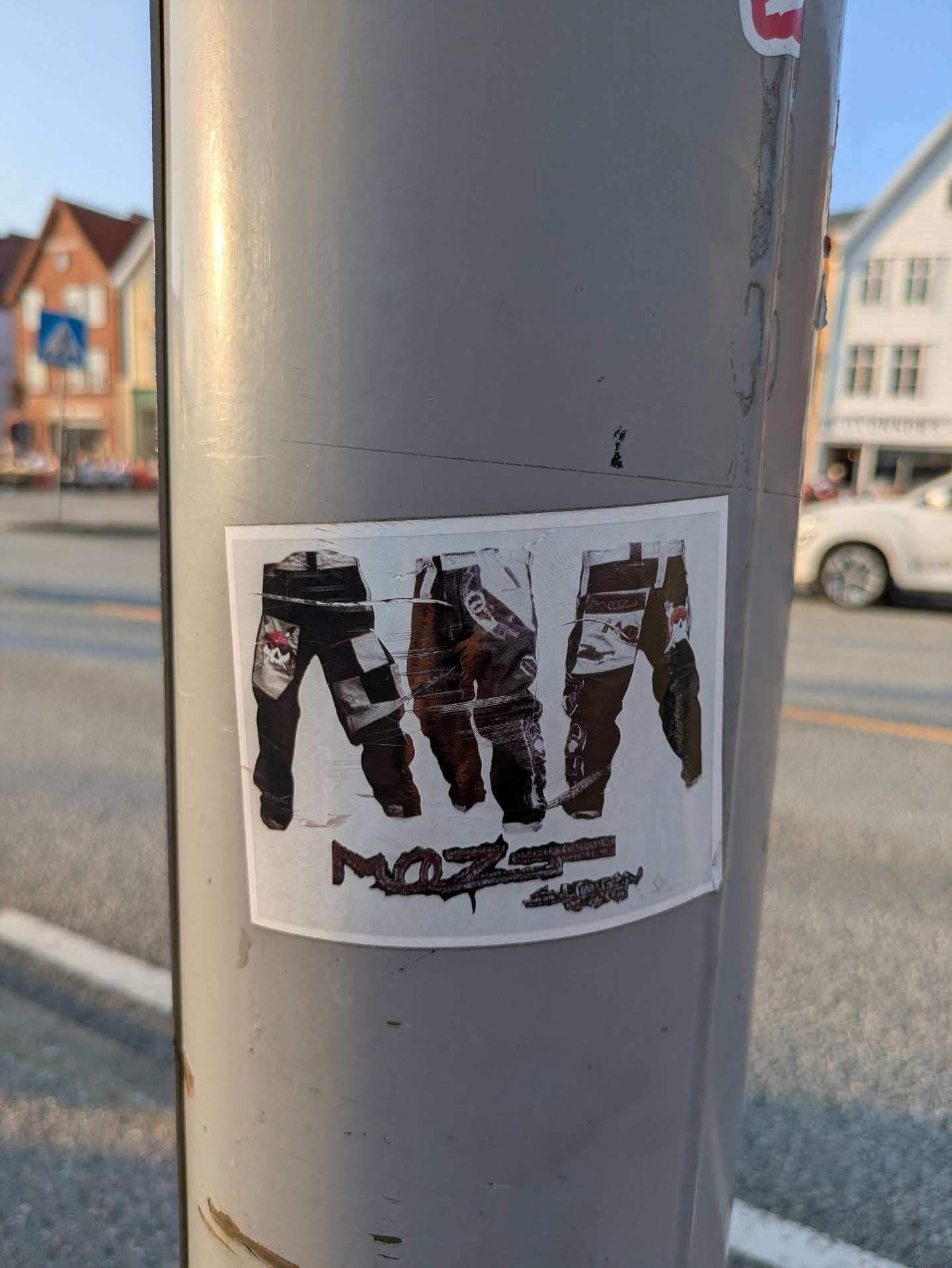 The sticker, placed on a grey utility pole, displays three distinct graphical representations of the lower halves of bodies wearing pants. The leftmost pair is black with white elements, including a checkerboard pattern and a patch with a pink skull and crossbones. The middle pair appears dark and worn, possibly brown or black. The rightmost pair is black with white detailing, including a small, possibly chicken-like character motif. Below these images, the word 'MOZZIE' is written in a distressed, stylized font, which is slightly worn. The sticker itself shows signs of wear, including creases and scratches.