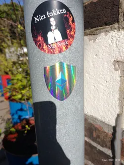 A holographic sticker featuring the Ethereum symbol within a shield shape.  The sticker is affixed to a metal pole alongside another sticker that says "Niet fokken Met Nouk" which translates from Dutch to "Don't fuck with Nouk".