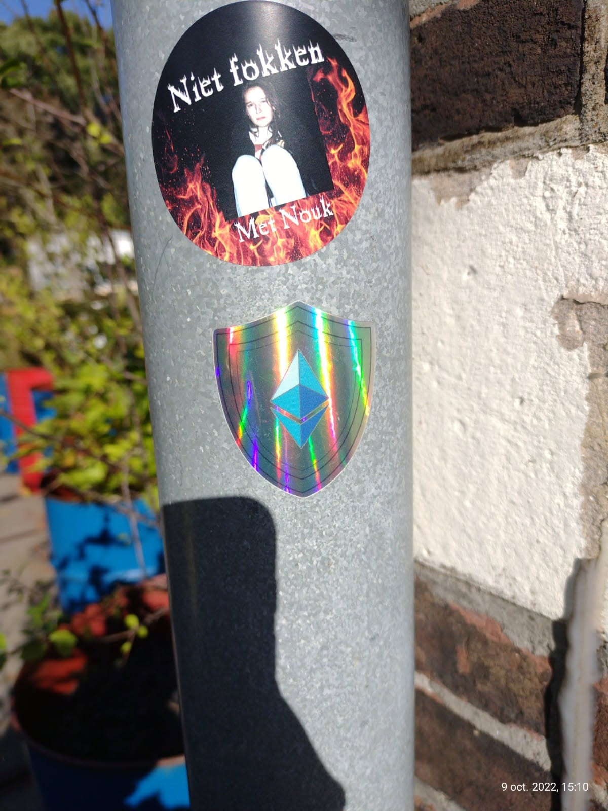 A holographic sticker featuring the Ethereum symbol within a shield shape.  The sticker is affixed to a metal pole alongside another sticker that says "Niet fokken Met Nouk" which translates from Dutch to "Don't fuck with Nouk".