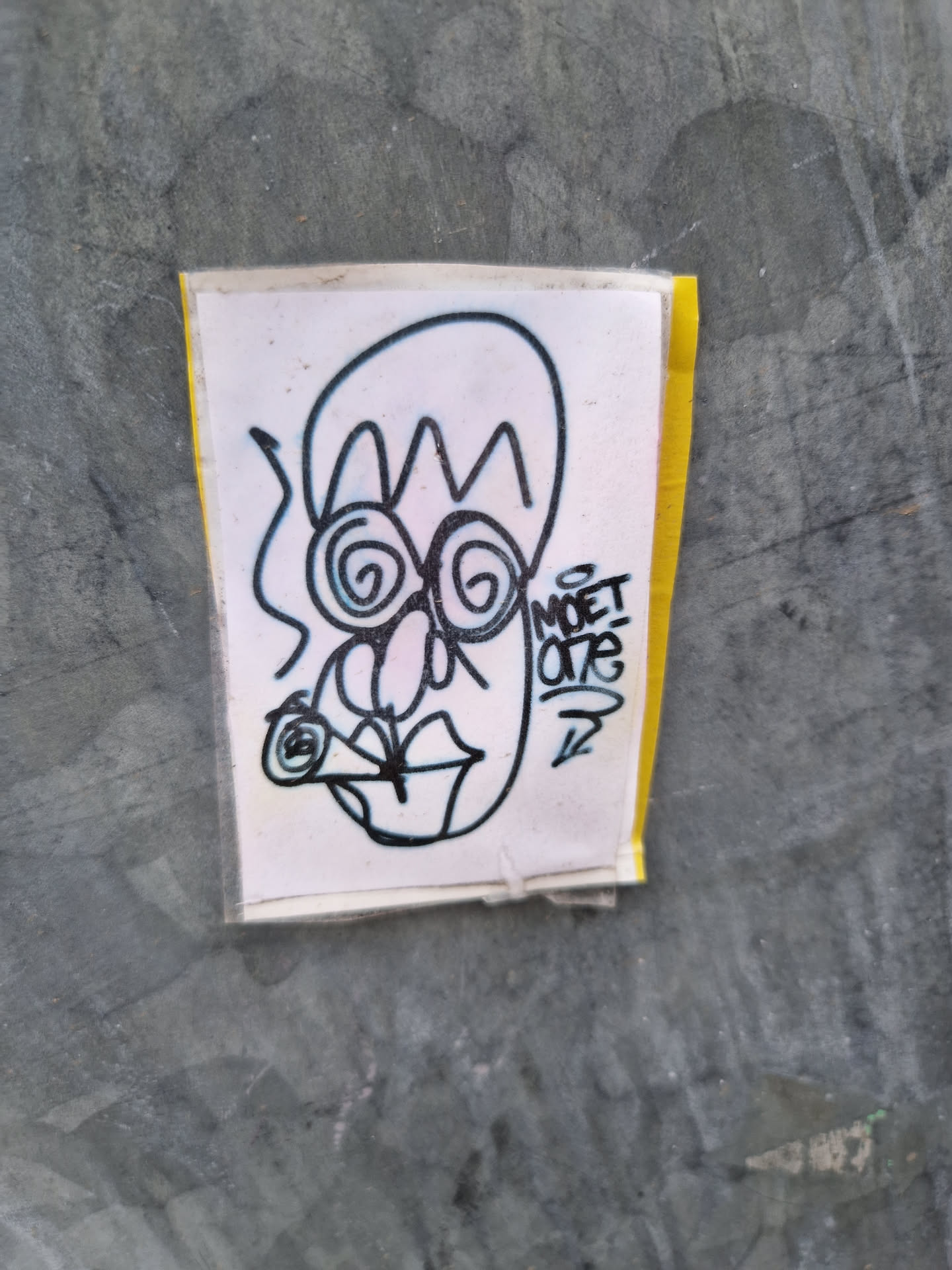 A hand-drawn sticker featuring a whimsical character with swirling eyes, a unique hairstyle, and what appears to be a cigarette in its mouth. The artist's tag, MOET ONE, is visible on the sticker.