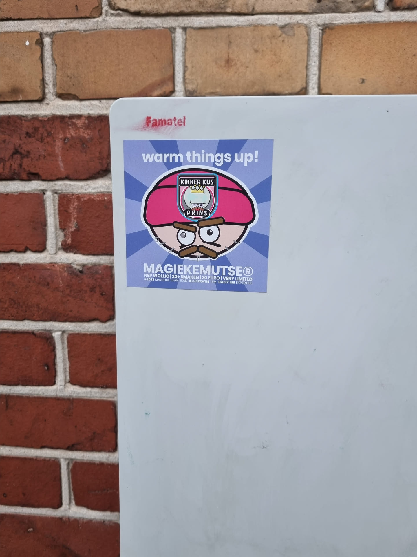 Sticker featuring a cartoon character with a pink hat and mustache. The text on the sticker says "MagiekeMutsen" and there's a logo that says "Knikker KUS DRINKS". The sticker is attached to a white board on a brick wall. There is also "Famatel" written on the board above the sticker.
