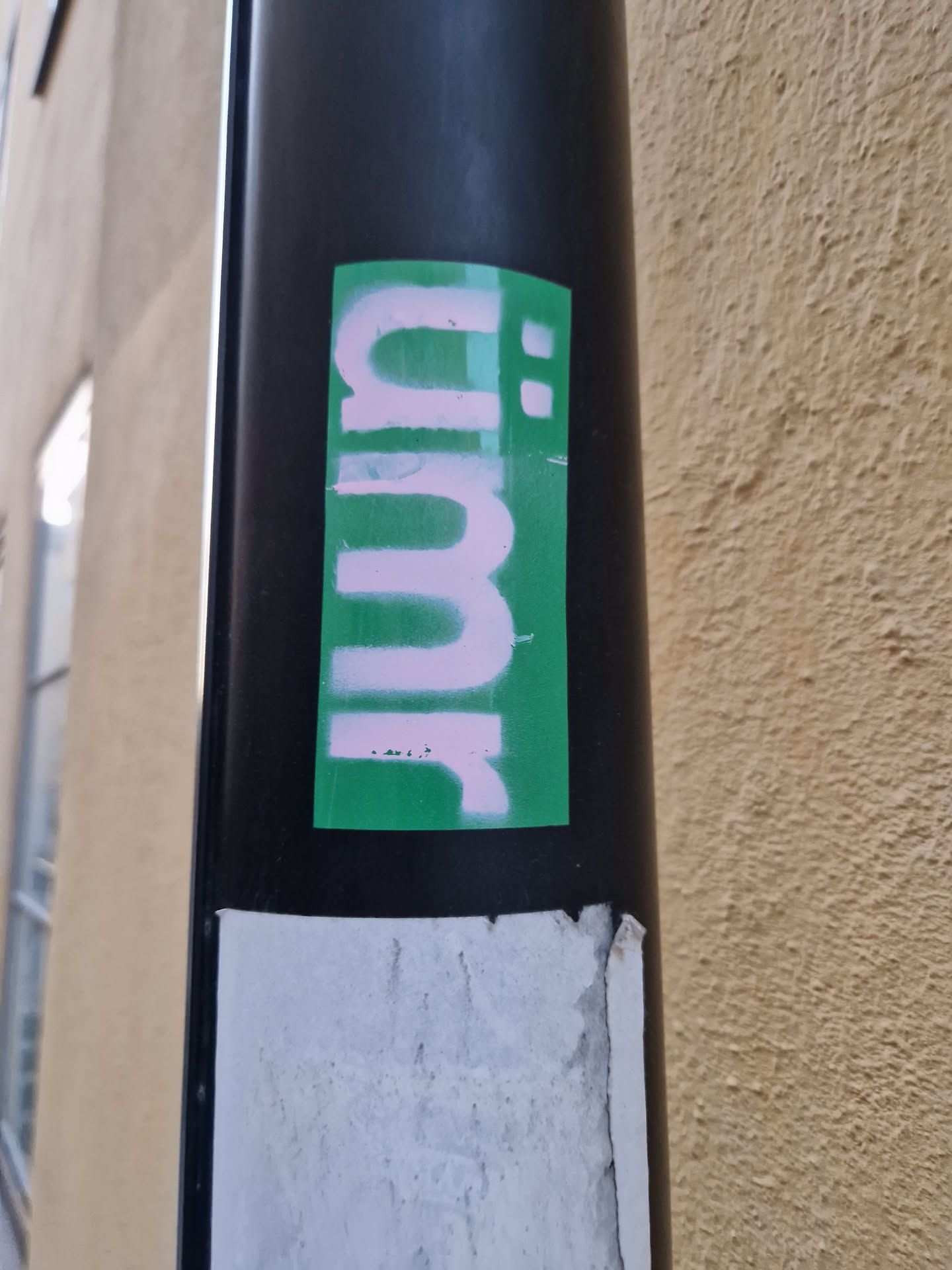 A green rectangular sticker with pink stylized lettering. The letters appear to be an abstract design rather than a recognizable word or phrase. The sticker is affixed to a black pole.