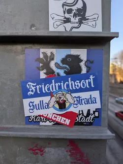 The central sticker cluster is composed of multiple layers affixed to a grey metal pole. The base layer is a rectangular sticker featuring a background of alternating vertical blue and white stripes. Superimposed on these stripes is a stylized black silhouette of a creature, possibly a lion or griffin, with a long, curled tail, clawed feet, and a single prominent eye. Overlaid on this is a prominent blue and white rectangular sticker. Its upper section displays 'Friedrichsort' in elegant white script on a blue background. Below this, a white band features a circular emblem of Popeye the Sailor, depicted flexing his bicep while holding a red bottle, with 'EST 1890' inscribed at the bottom. To the right of Popeye, 'Sulla Strada' is written in a similar ornate white script on a blue background. Partially visible beneath these layers is a small red sticker with white text, which reads 'Karu' and 'NCHEN Stadt', likely indicating 'München Stadt'.