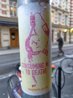 A sticker featuring an image of a person being hanged with the text 'CONSUMING ME TO DEATH!' and a website address. The style is simple, with a distressed look. The sticker is attached to a metal pole on a city street.