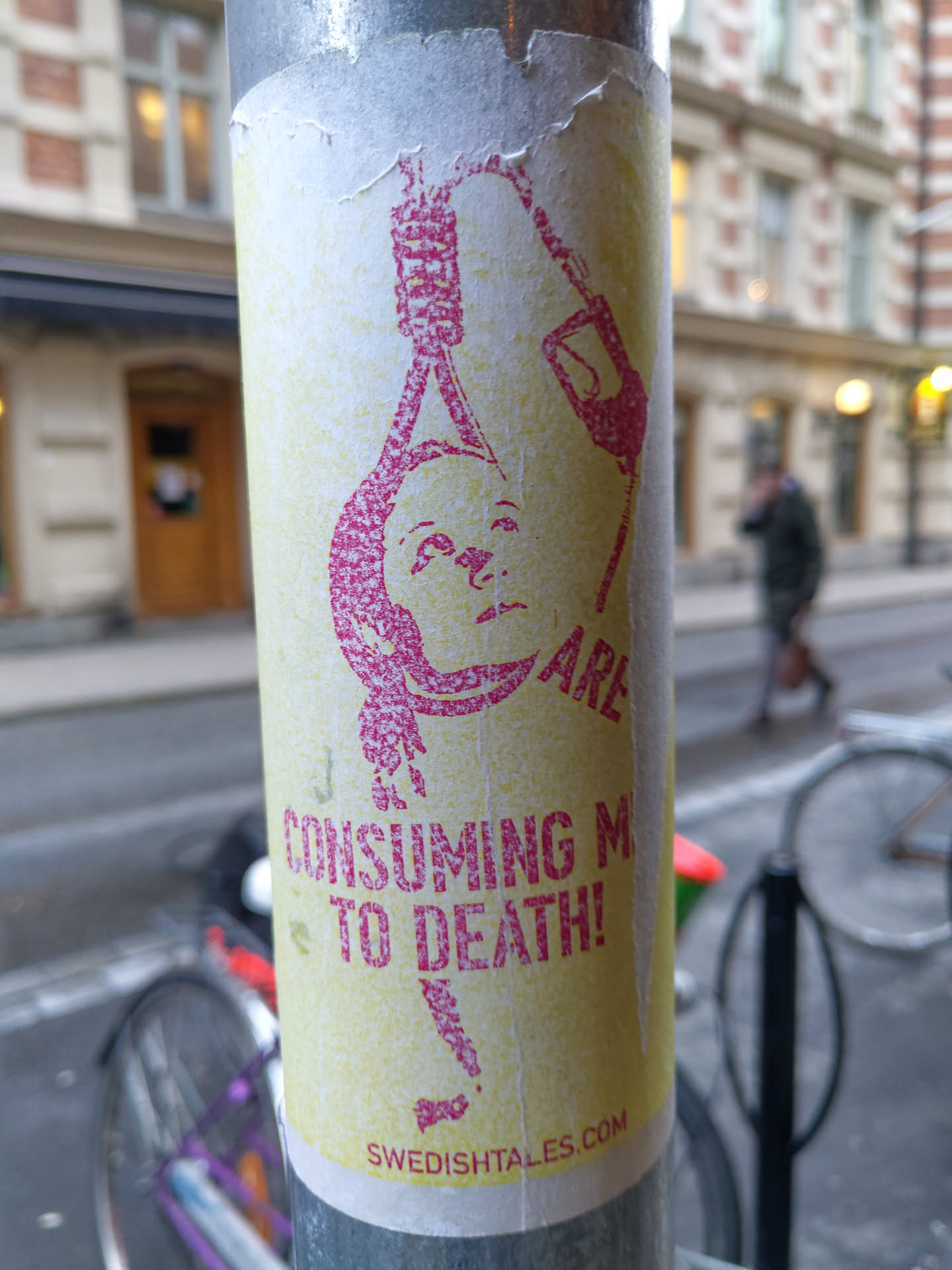 A sticker featuring an image of a person being hanged with the text 'CONSUMING ME TO DEATH!' and a website address. The style is simple, with a distressed look. The sticker is attached to a metal pole on a city street.