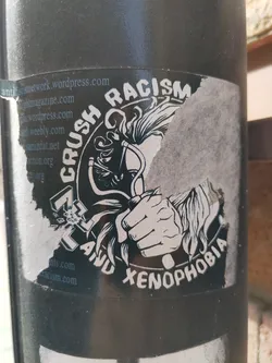 A sticker with a graphic design depicting a hand crushing symbols of racism and xenophobia.  The sticker is partially damaged.