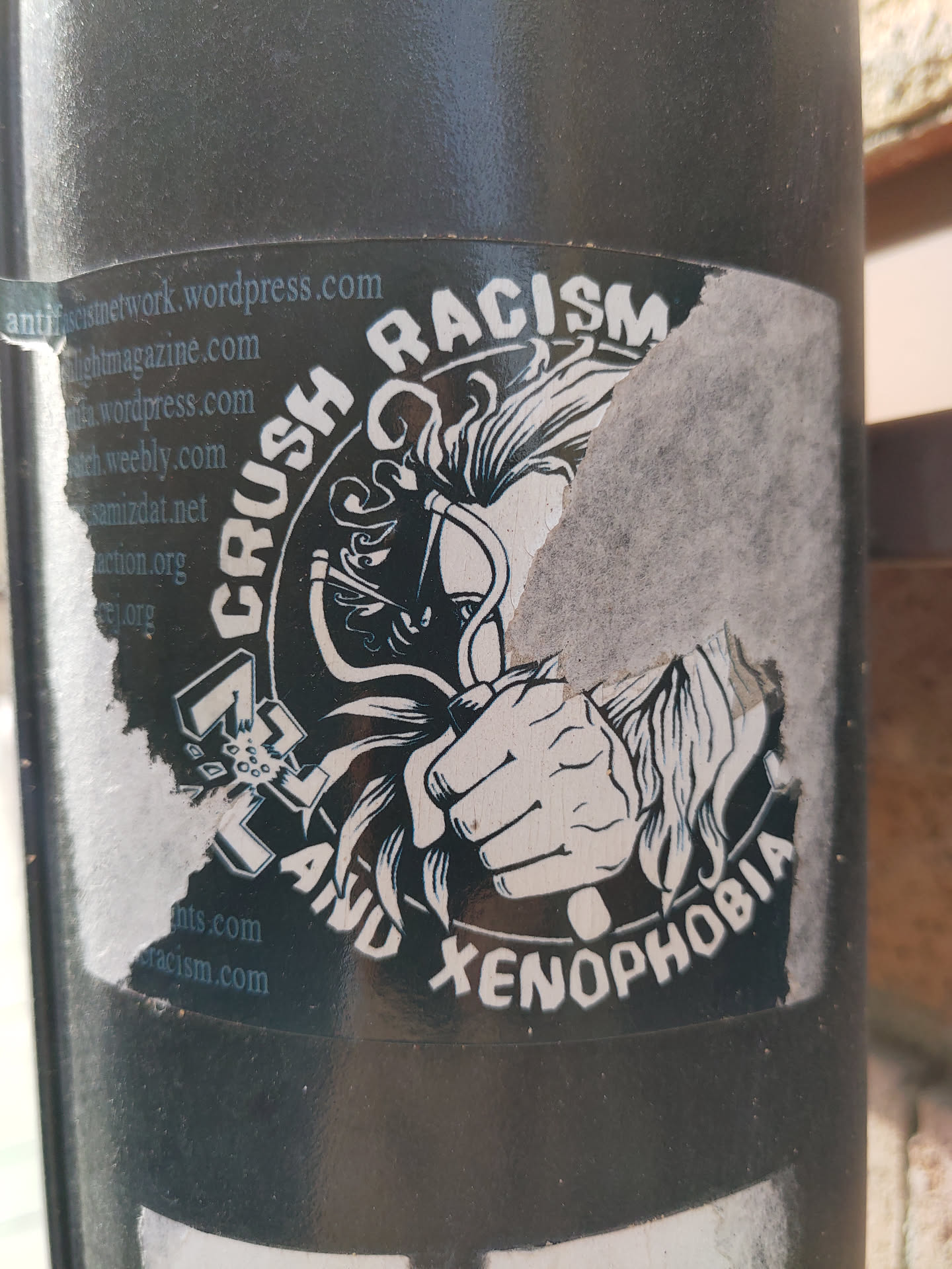 A sticker with a graphic design depicting a hand crushing symbols of racism and xenophobia.  The sticker is partially damaged.