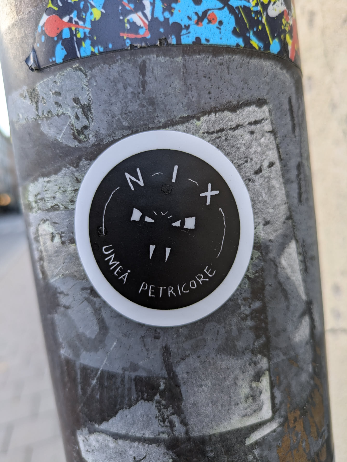 Round black and white sticker with a minimalist design. It features a stylized face-like image with N-I+ symbols and the words UMEÄ and PETRICORE.
