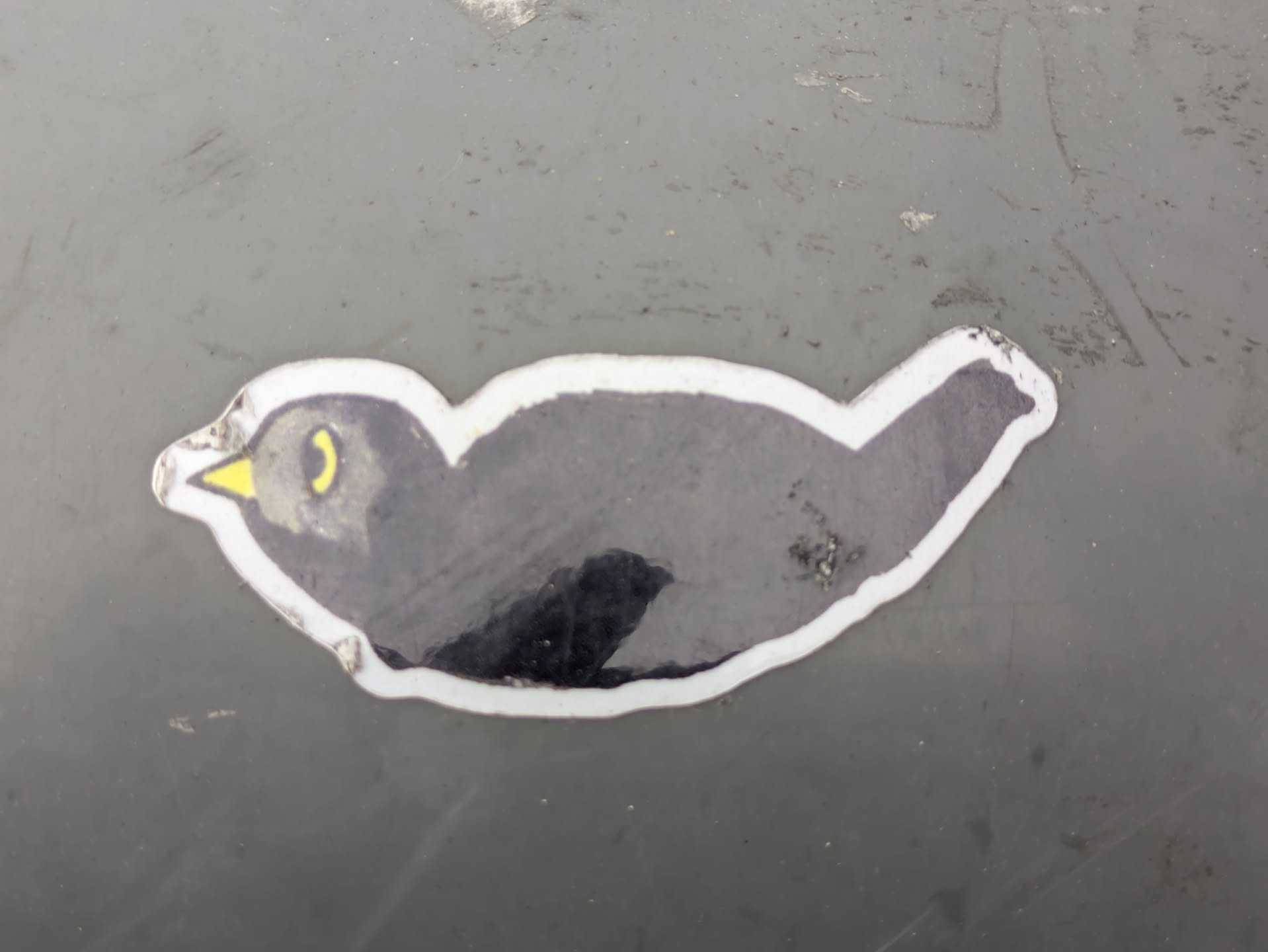 A small sticker featuring a simple drawing of a dark-colored bird with a yellow beak. The bird is depicted in a side profile, and its body is slightly curved. The sticker is affixed to a dark grey surface.