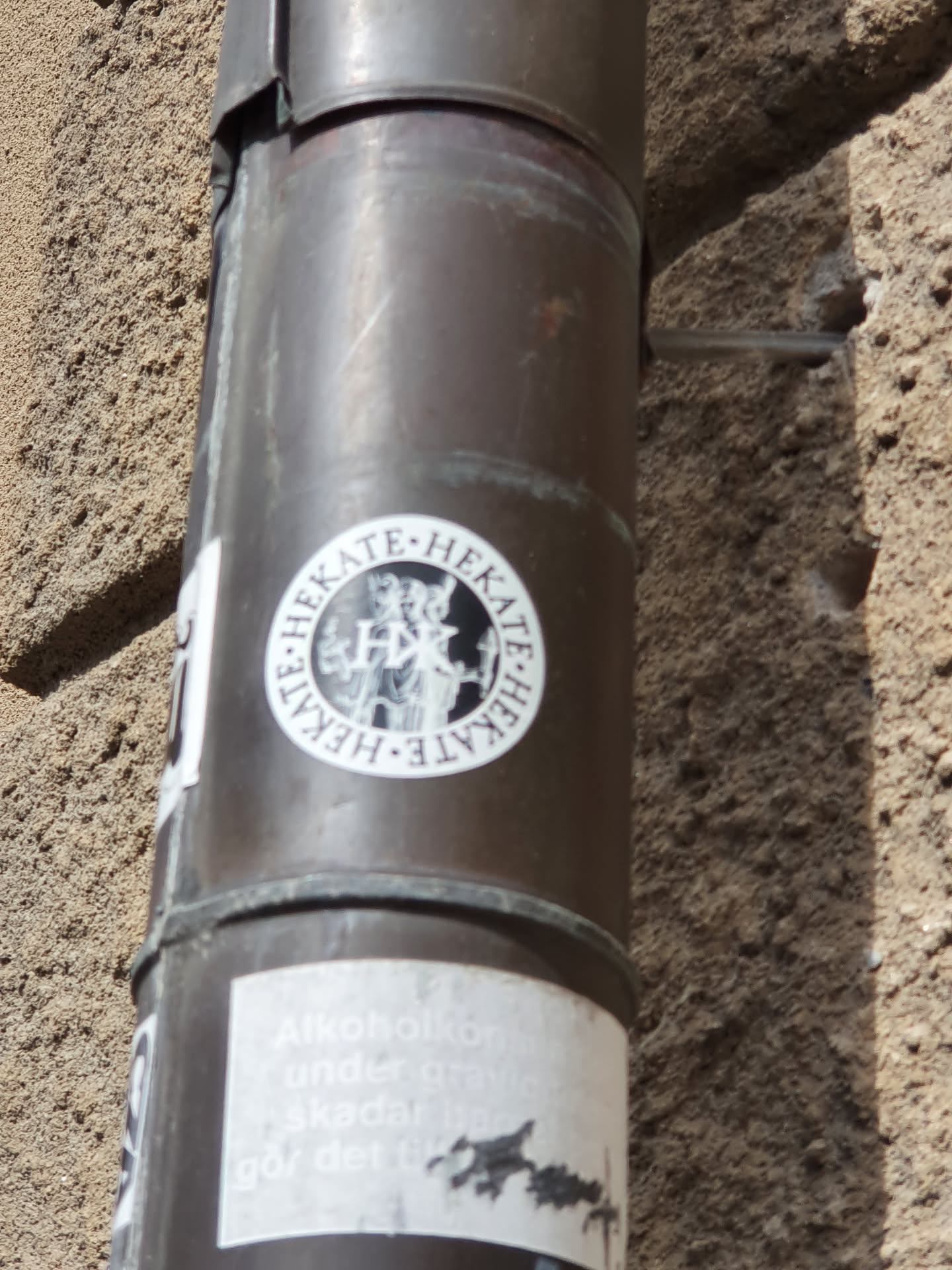 A round sticker featuring the name HEKATE multiple times and an image of the goddess in the center is affixed to a metal downspout. A second sticker below is partially visible, but illegible.