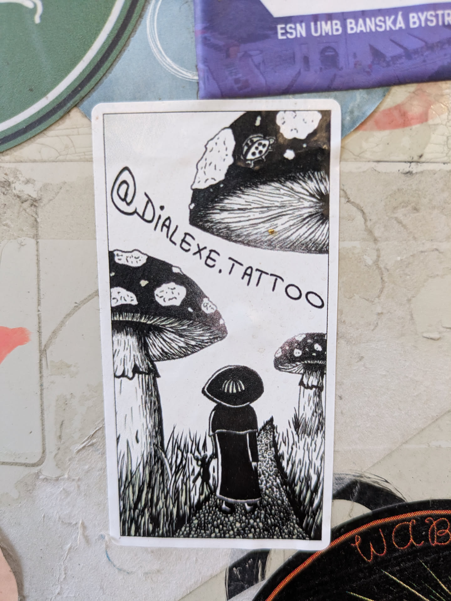 A black and white sticker features a person standing in a mushroom forest. The person is wearing a wide brimmed hat. The sticker features the instagram handle @dialexe.tattoo.