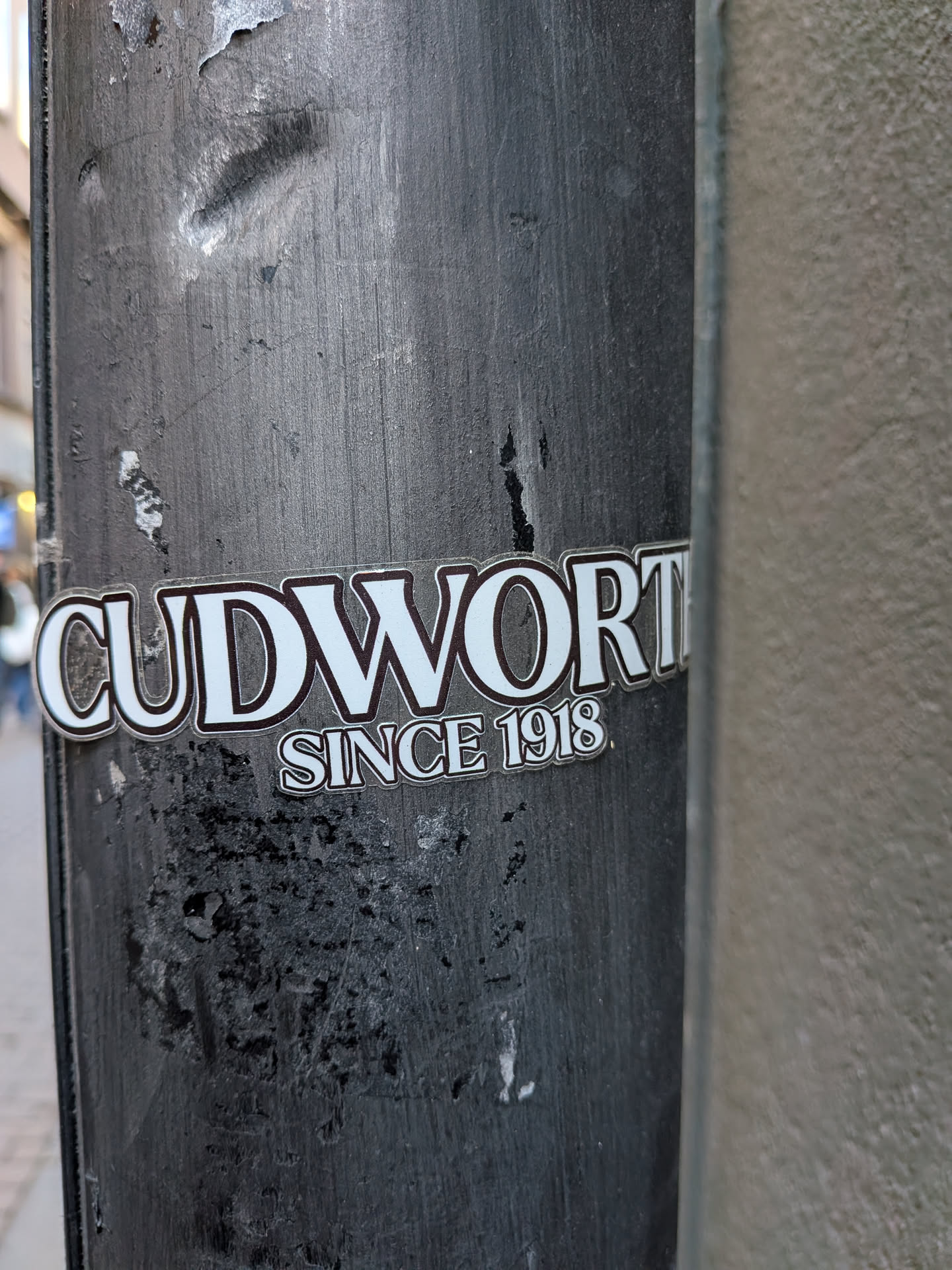 A contour-cut sticker featuring the word 'CUDWORTH' in large, capitalized white serif lettering with a dark outline. Beneath the right side of the main word, the phrase 'SINCE 1918' is printed in a matching outlined serif typeface. The sticker is adhered to a weathered, dark painted vertical pole.