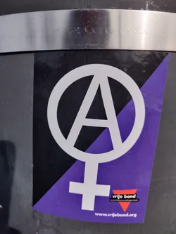 The sticker shows an anarchist symbol combined with a female gender symbol, overlaid on a purple and black background.  It includes a small logo and website address at the bottom.
