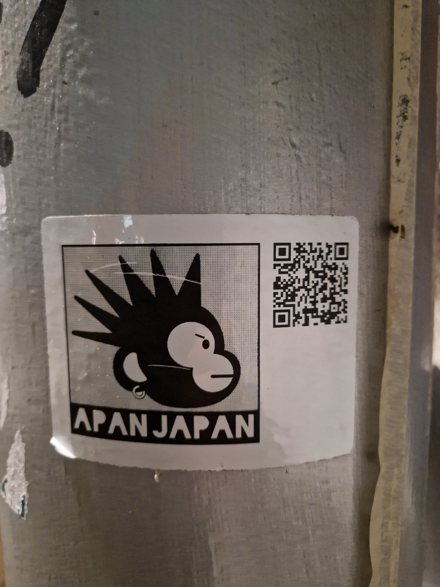 A black and white sticker featuring a monkey with spiky hair, a piercing, and an angry expression.  The text 'APANJAPAN' is displayed below the monkey. A QR code is present on the right side of the sticker.