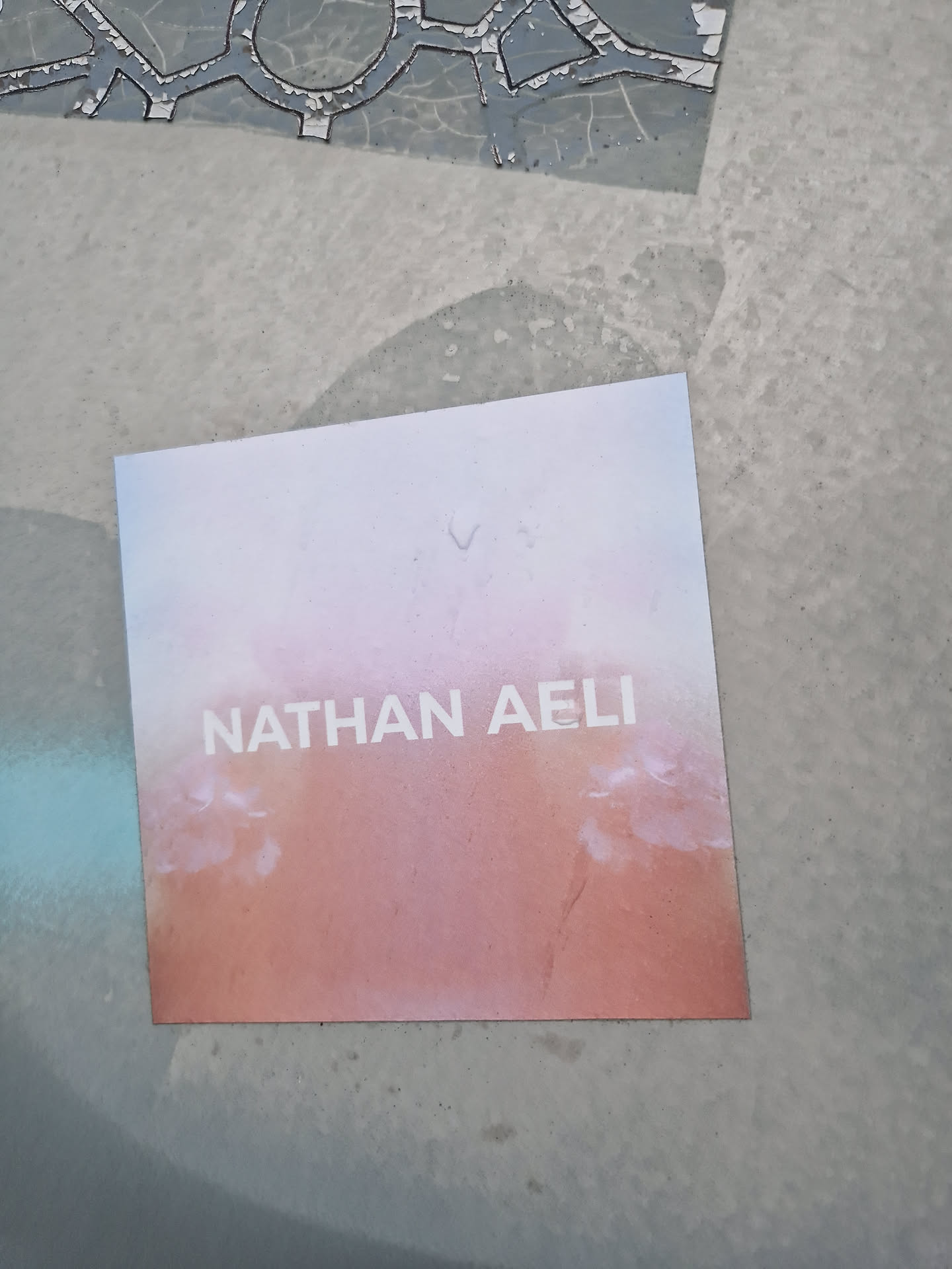 A square sticker with a peach and white gradient background and the name NATHAN AELI in sans-serif font. The sticker is placed on a gray surface.