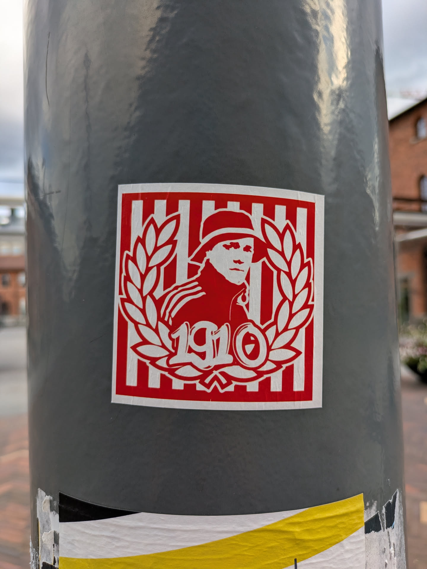 The sticker is square with a thin white border. The main background is composed of vertical red and white stripes. Centered on this striped background is a white silhouette of a person, likely a man, wearing a hat, facing slightly to the right. This figure is framed by a white laurel wreath, open at the top. Below the figure, within the wreath, are the stylized numbers "1910" in white.