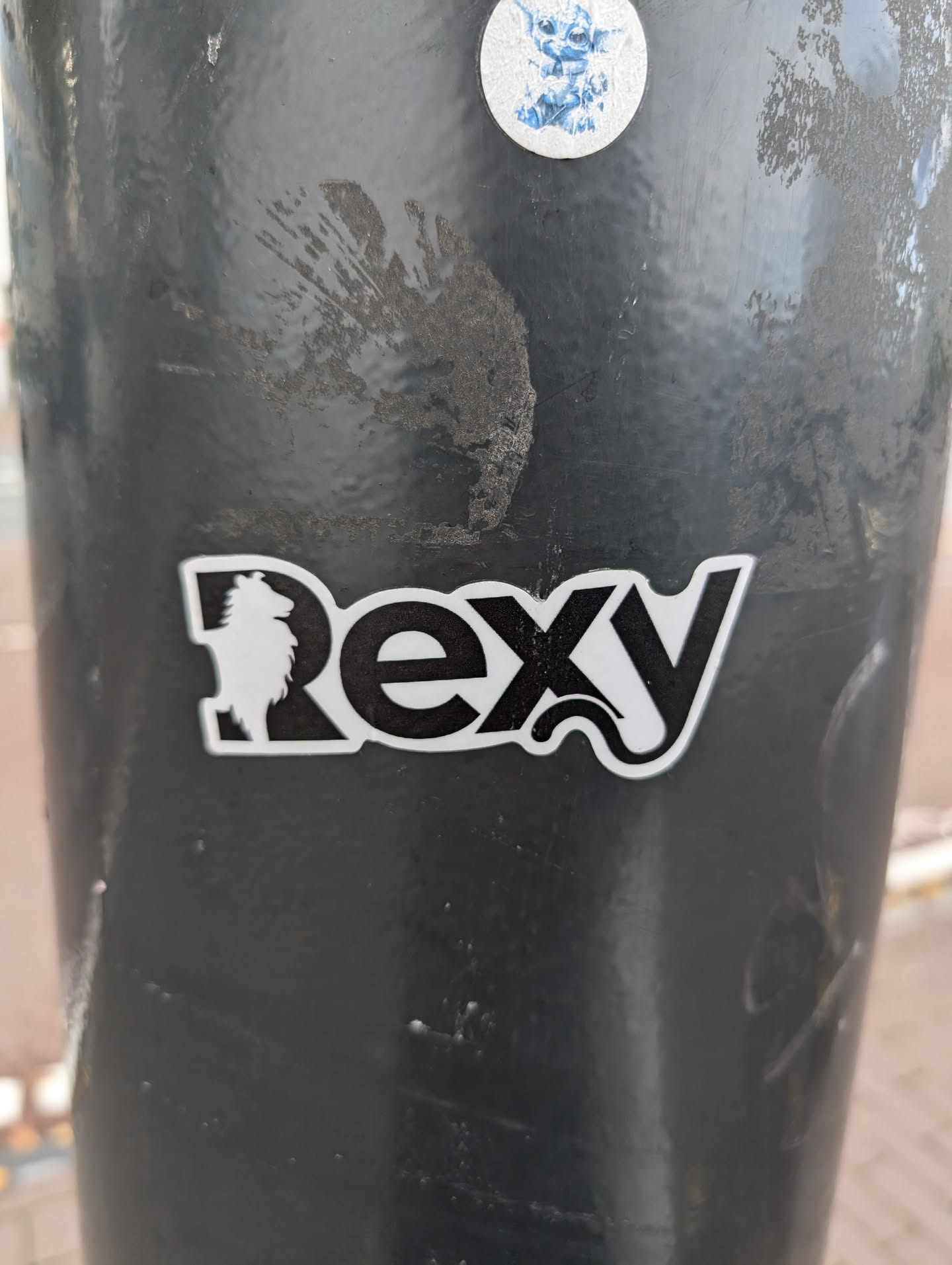 Black and white sticker with the word Rexy and a small dog-like figure. The sticker is placed on a dark gray pole.