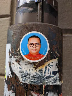 A circular sticker featuring a portrait of a man with a light brown complexion, short dark hair, and round, dark-rimmed glasses. He is wearing an orange or saffron-colored robe. The background of the portrait is a bright, solid blue. The sticker has a thin white border around the blue circle. Thai script is written in white along the curved edges of the blue background: 'พุทธรักษา' (Buddha protects) on the left, 'ธัมมังรักษา' (Dhamma protects) at the bottom, and 'สังฆังรักษา' (Sangha protects) on the right. The sticker is affixed to a heavily rusted metal pole, with some wear visible around its edges.