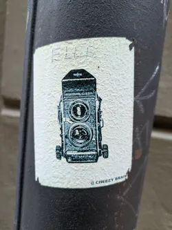 A sticker featuring a drawing of a vintage Rolleiflex camera. The sticker is off-white with teal-colored illustration and has the text "© CHEEZY BRAIN" at the bottom.