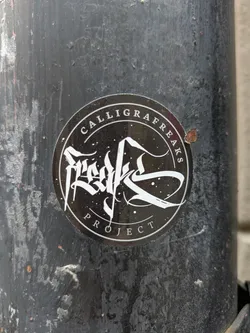 Round black and white sticker with the text "Calligrafreaks Project" and a stylized calligraphy design in the center. The sticker is affixed to a dark gray, weathered surface.