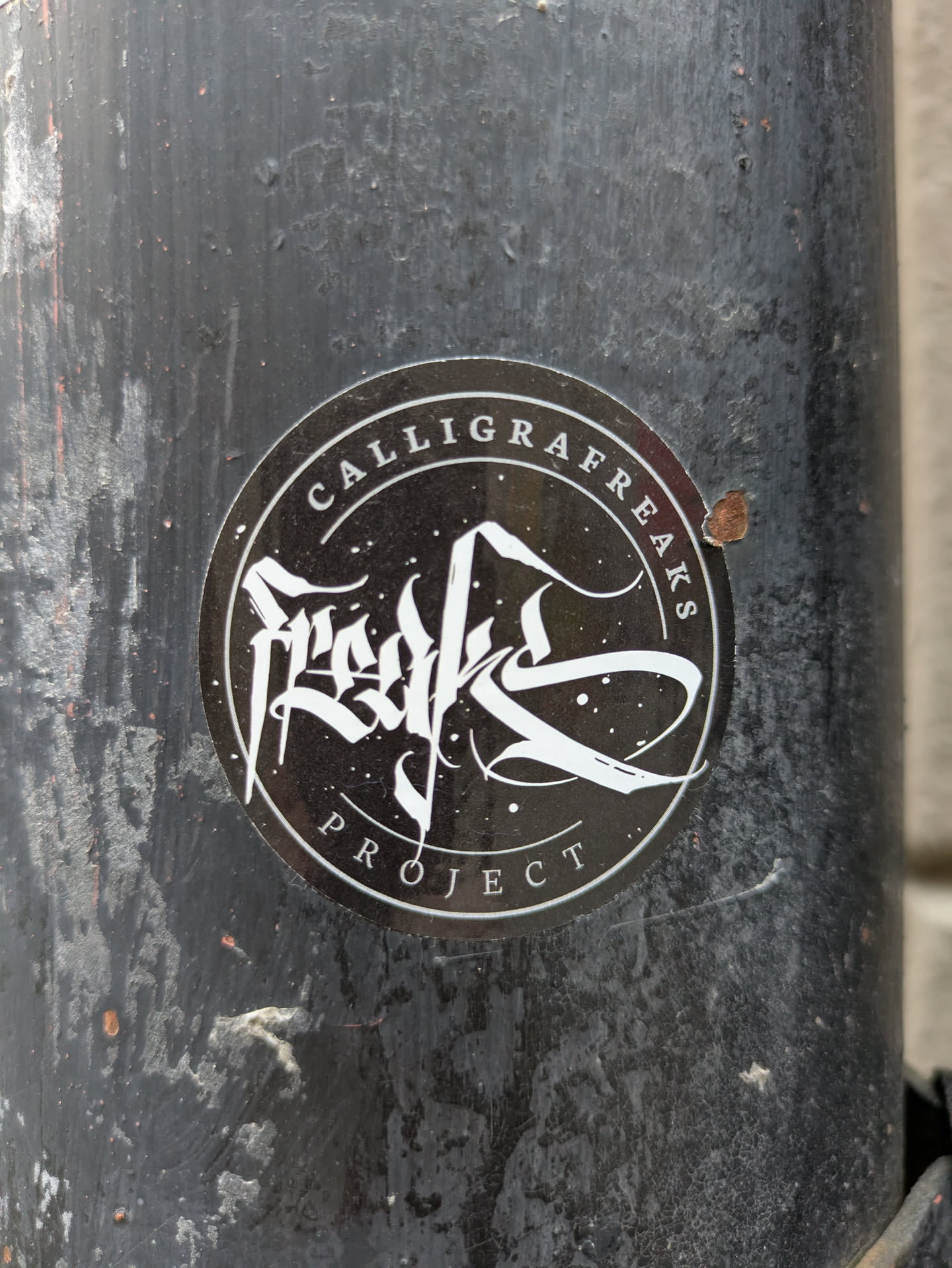 Round black and white sticker with the text "Calligrafreaks Project" and a stylized calligraphy design in the center. The sticker is affixed to a dark gray, weathered surface.