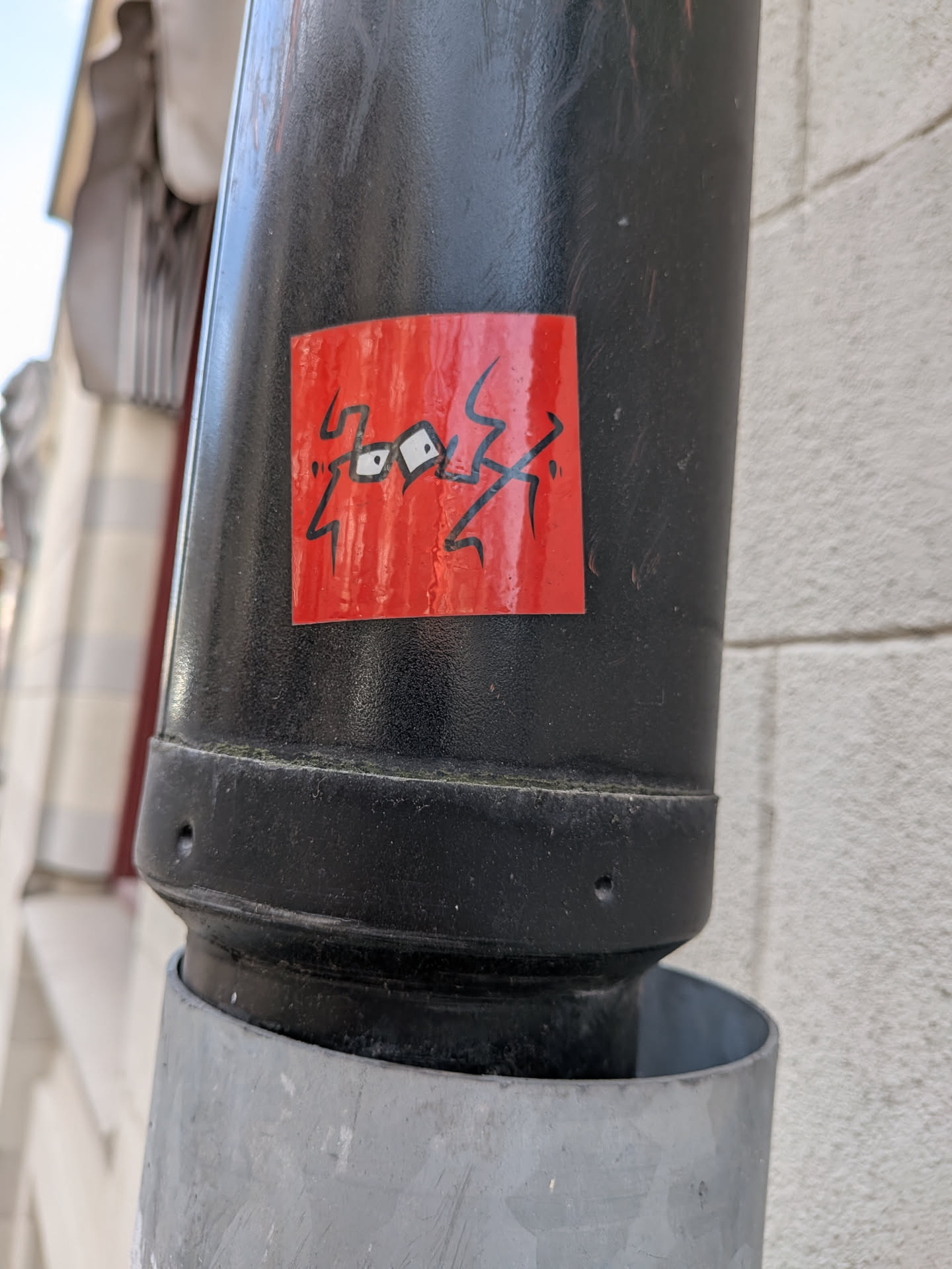 The sticker is rectangular with a vibrant, slightly textured red background. It features a bold, abstract design in black. The central elements are two white-outlined black shapes that strongly resemble stylized eyes, connected by a thin black line. Surrounding these 'eyes' are jagged, angular black lines that extend outwards, giving the impression of lightning bolts, energy, or a chaotic scribble. The overall aesthetic is graphic and minimalist, relying on strong color contrast and dynamic lines.