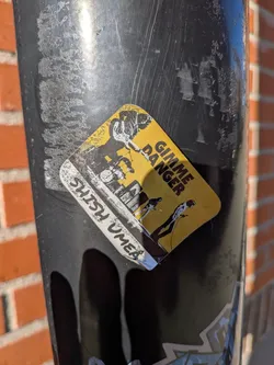 The sticker is roughly diamond-shaped with rounded corners, predominantly yellow with black and white graphics. The top portion features a black and white illustration of what appears to be a band playing instruments, possibly a drummer and guitarists, with a gritty, hand-drawn aesthetic. To the right of the illustration, in bold black sans-serif capital letters, is the text 'GIMME DANGER'. Below the main illustration, a white band with black text reads 'SHISH OMEA'. The sticker shows some signs of wear and tear, with slight peeling at the edges and some scuffing on the surface, indicating it has been applied for some time.