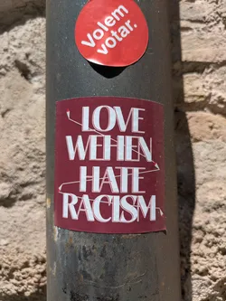 A rectangular sticker on a gray pole, next to a brick wall. The sticker has a burgundy background with white text that says 'Love When Hate Racism'. There are decorative lines around the text, giving it a hand-drawn feel. Above the rectangular sticker there is a round sticker that says 'Volem votar'.