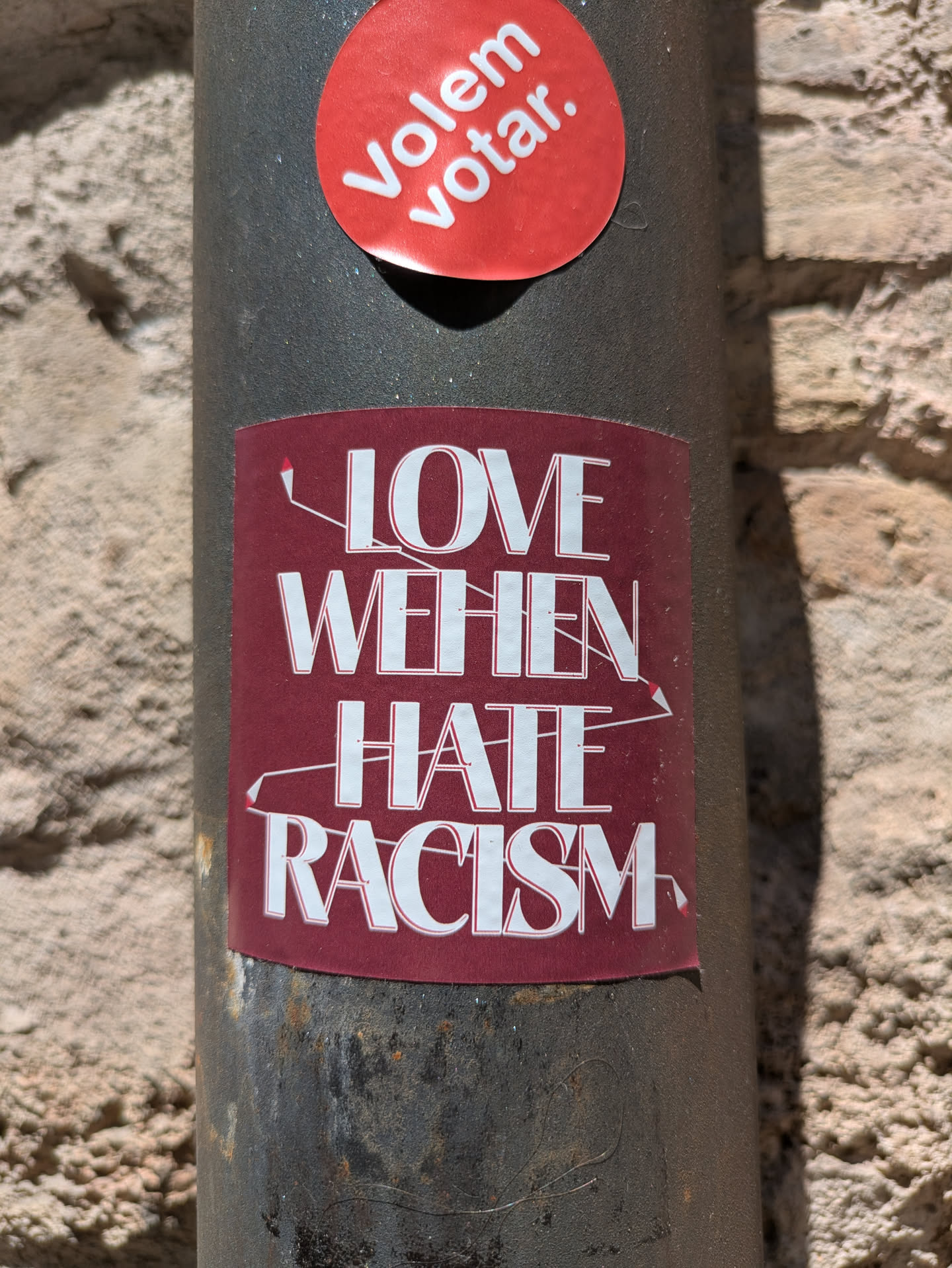 A rectangular sticker on a gray pole, next to a brick wall. The sticker has a burgundy background with white text that says 'Love When Hate Racism'. There are decorative lines around the text, giving it a hand-drawn feel. Above the rectangular sticker there is a round sticker that says 'Volem votar'.
