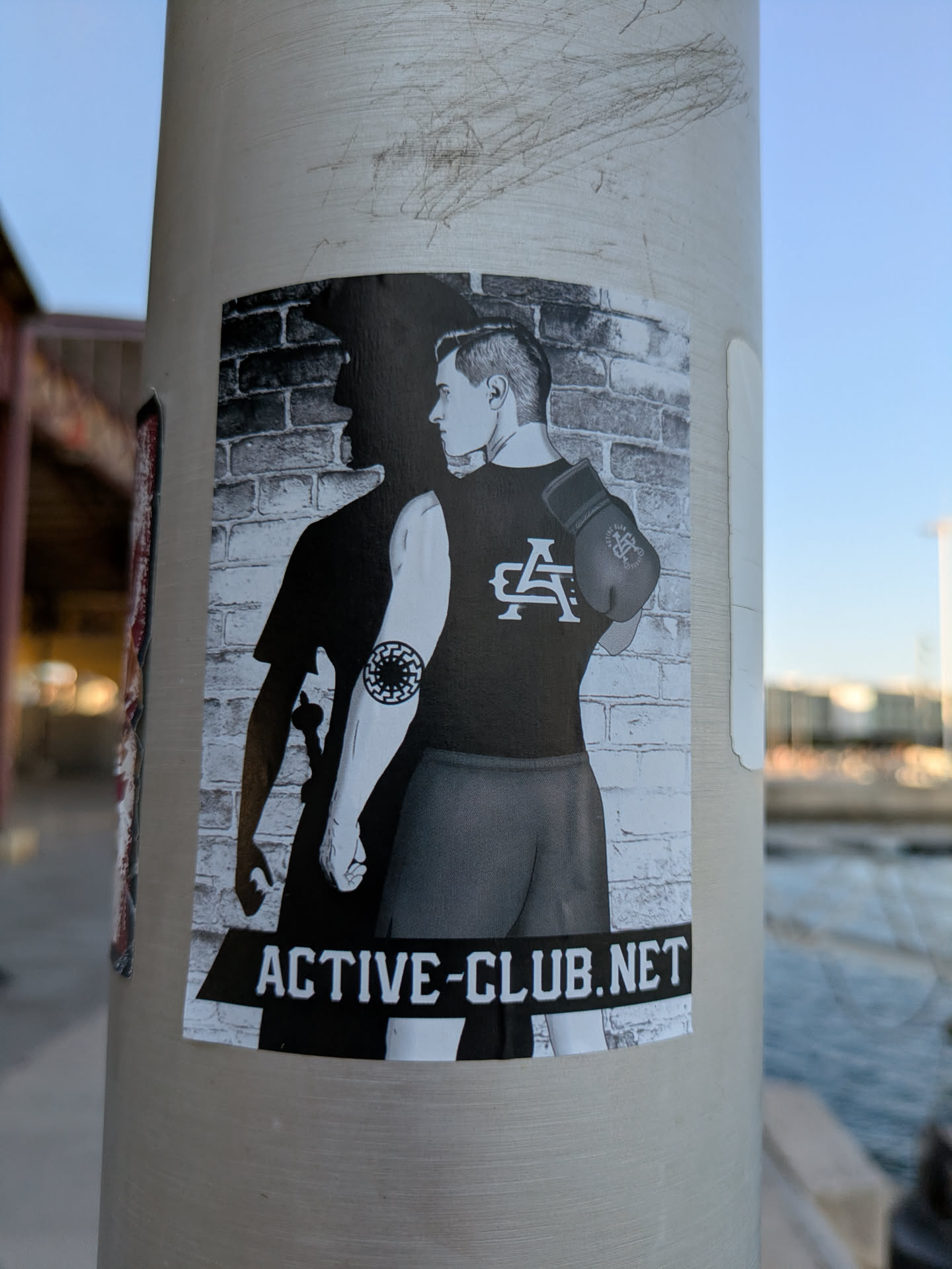 The sticker is black and white and shows a man standing in front of a brick wall with a shadow behind him. He is wearing a tank top with two overlapping letters on it, shorts, and a boxing glove on his shoulder. He has a tattoo on his bicep. The text ACTIVE-CLUB.NET is at the bottom of the sticker.